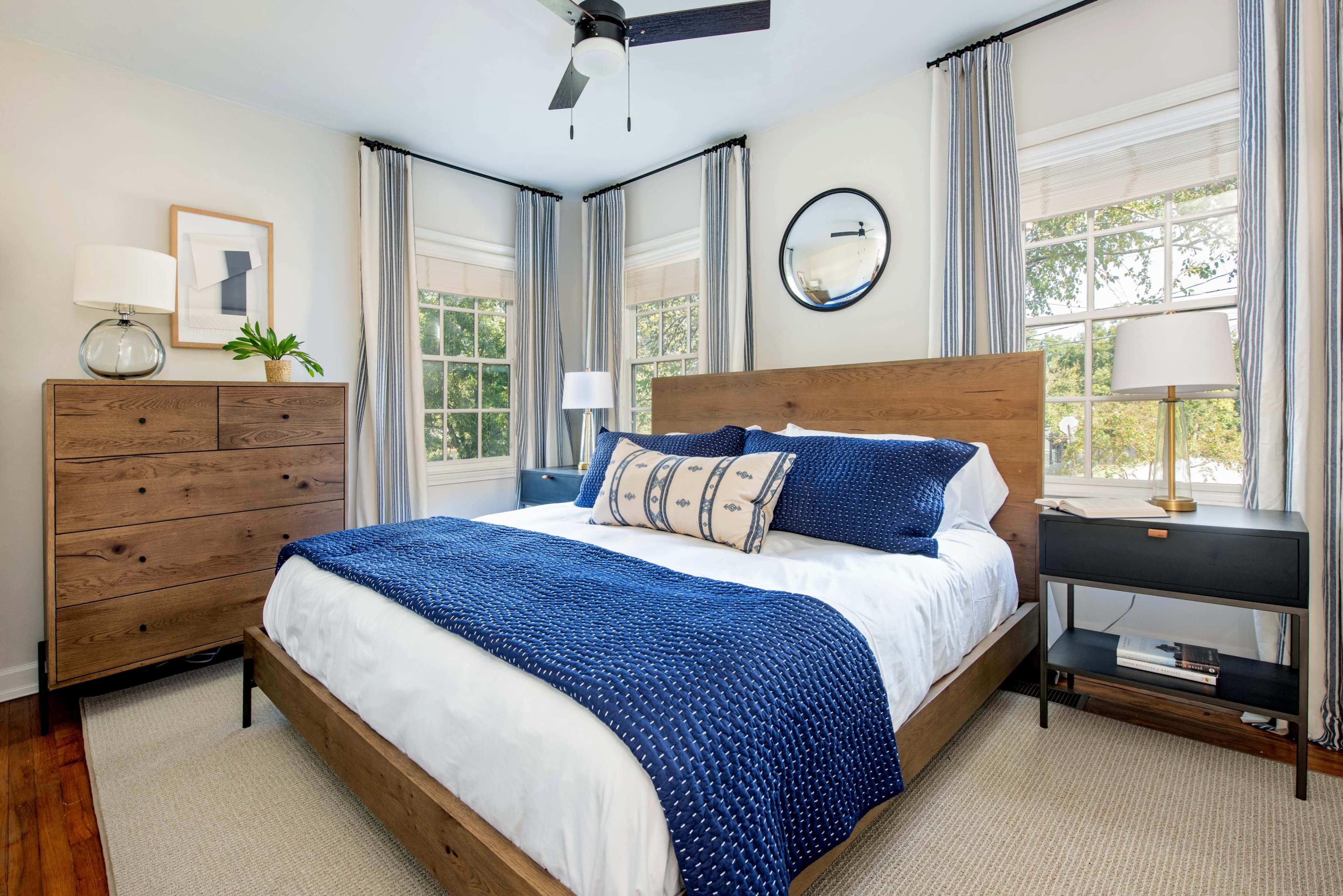 A neatly arranged bedroom with a wooden bed, blue and white bedding, a dresser, and large windows allowing natural light to enter.