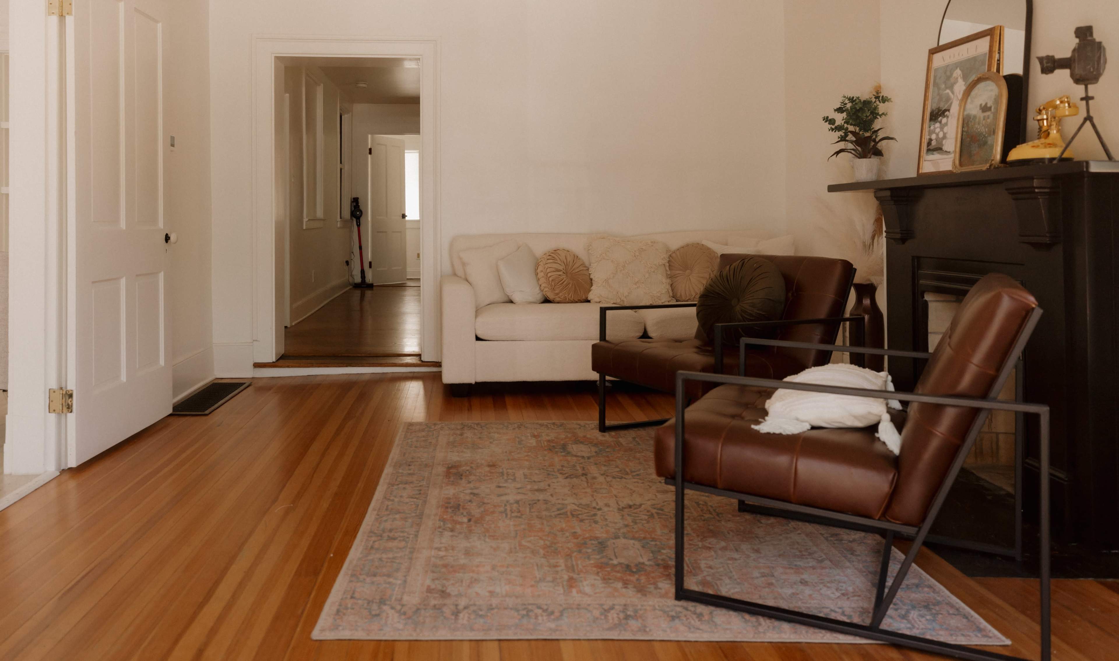 The image shows a living room with a beige sofa, a brown leather armchair, and a patterned area rug on hardwood flooring, with a fireplace and doorways leading to other rooms.