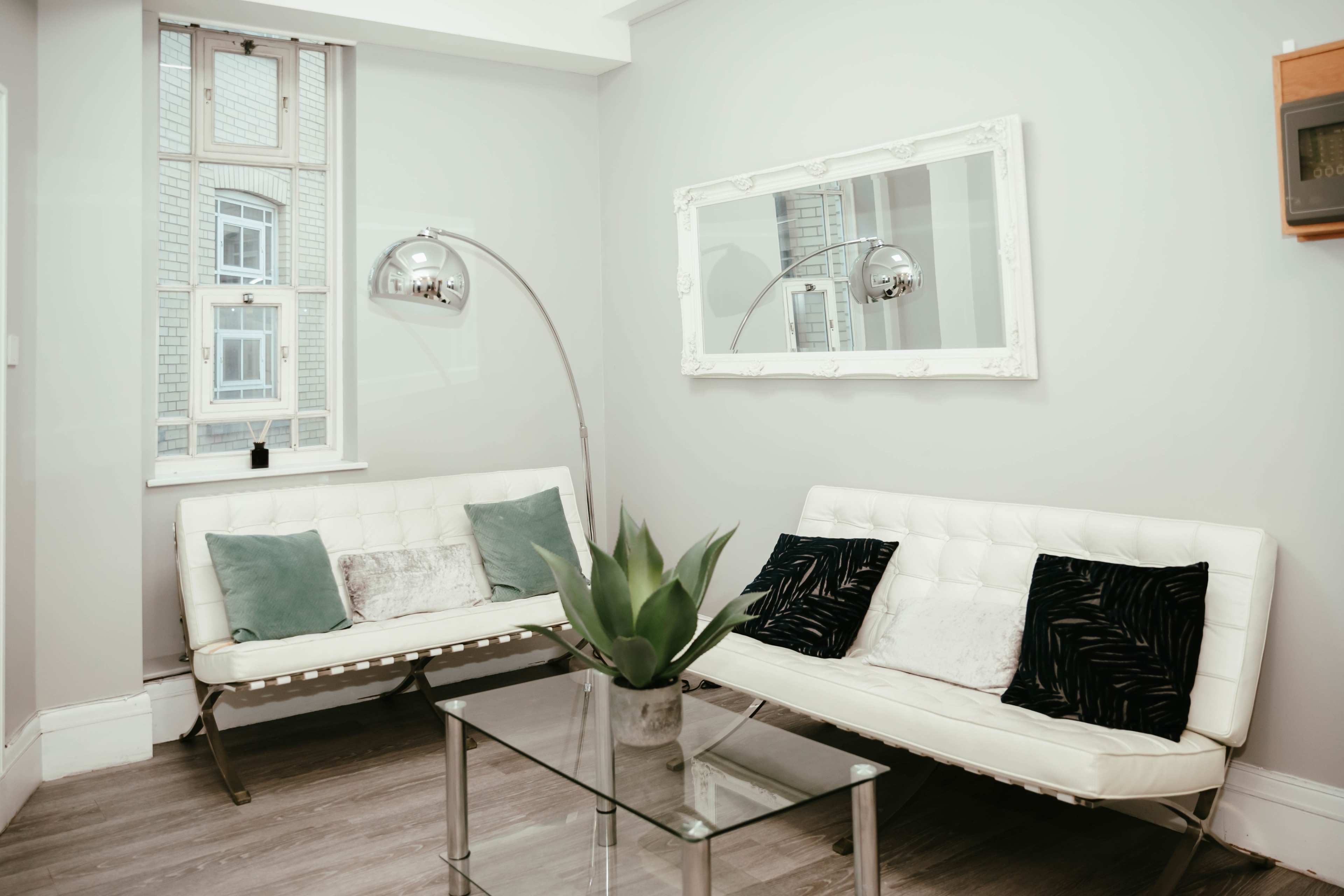 A modern seating area features two white sofas with cushions, a glass coffee table, and a floor lamp, set against a light gray wall.