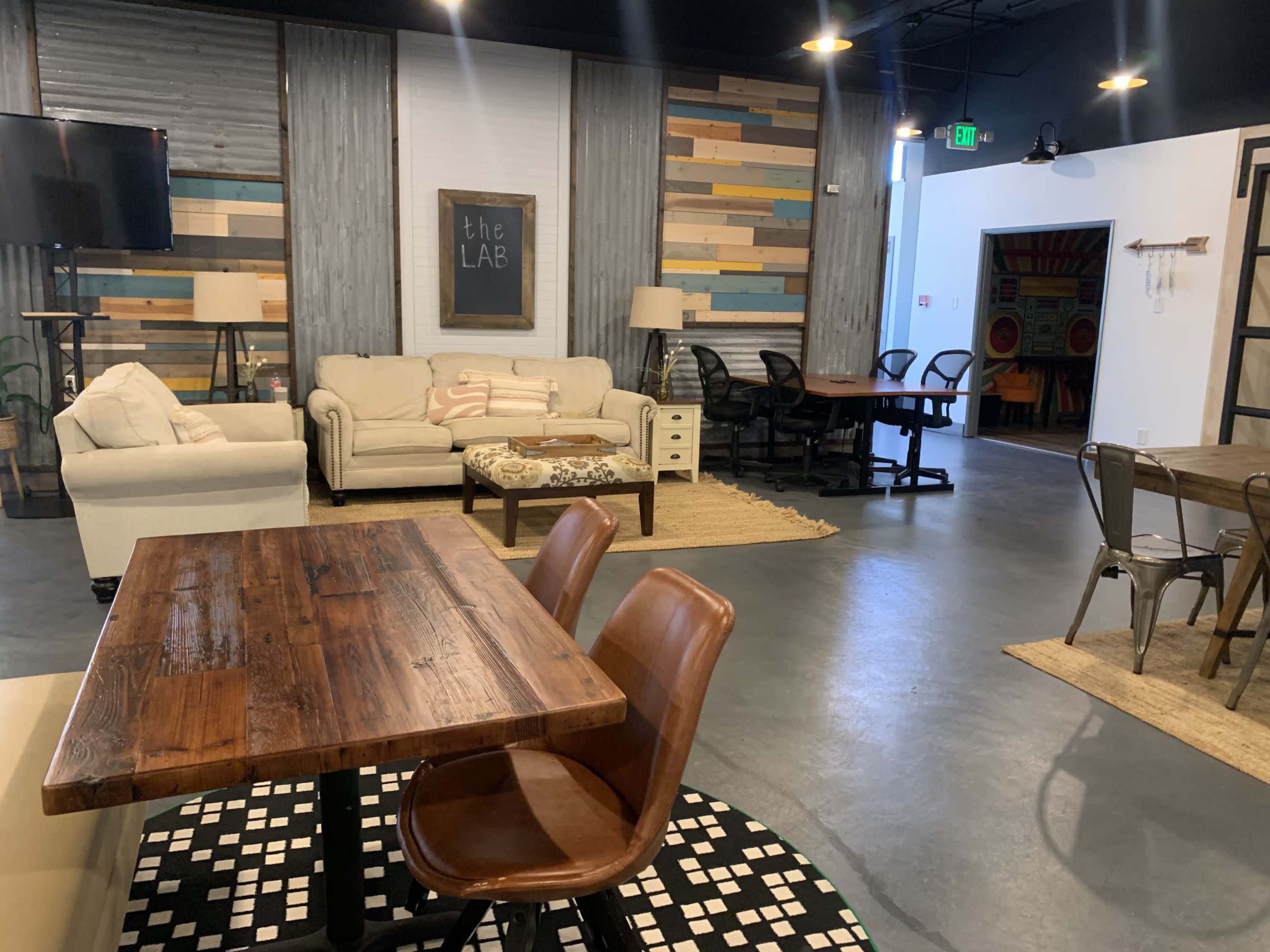 The image shows a spacious, modern coworking area with a mix of seating arrangements, including a wooden table and chairs, a couch, and colorful wall panels.
