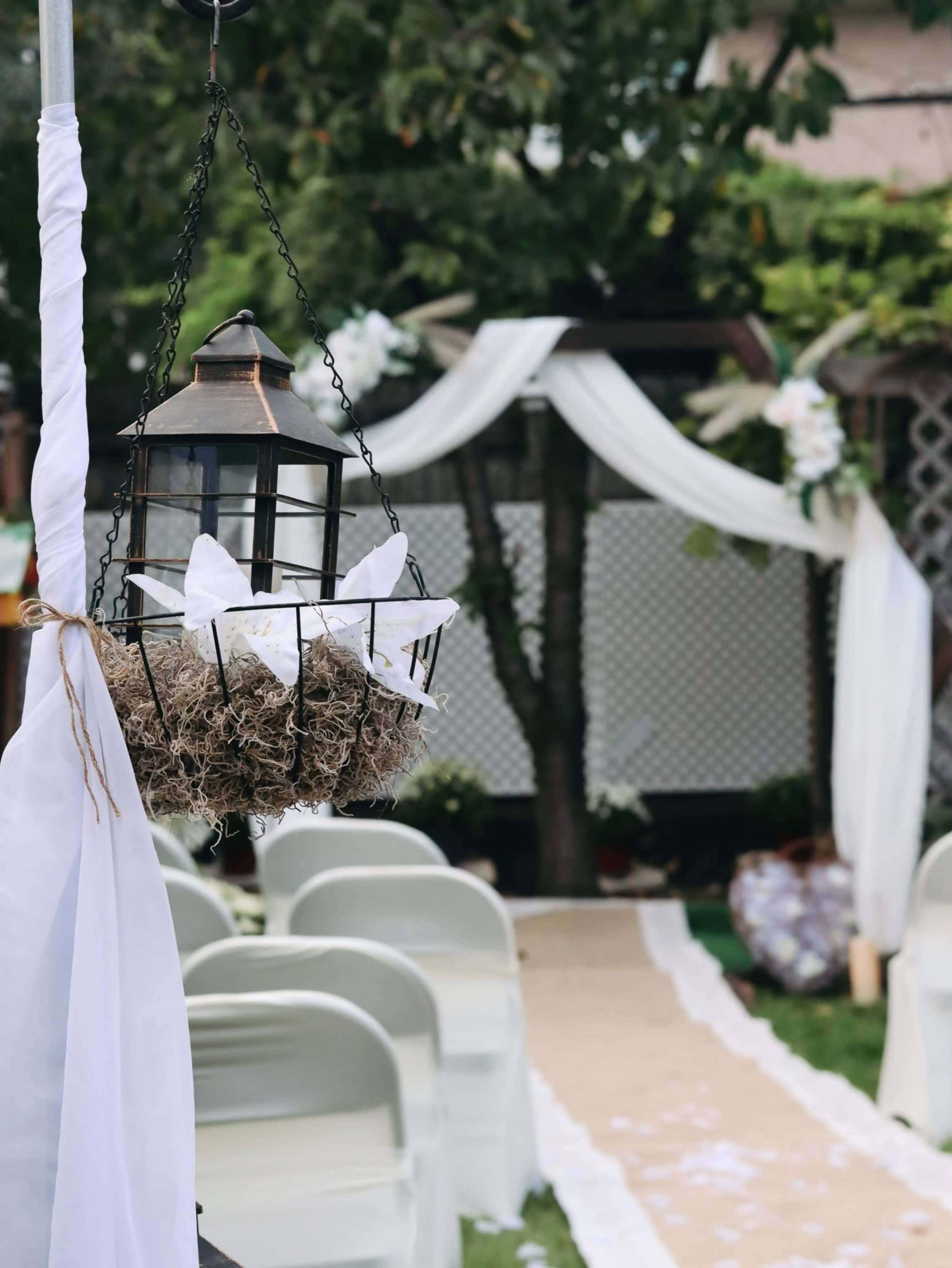 A decoratively arranged outdoor wedding venue with chairs facing a center aisle, flanked by a floral arch and a hanging lantern.