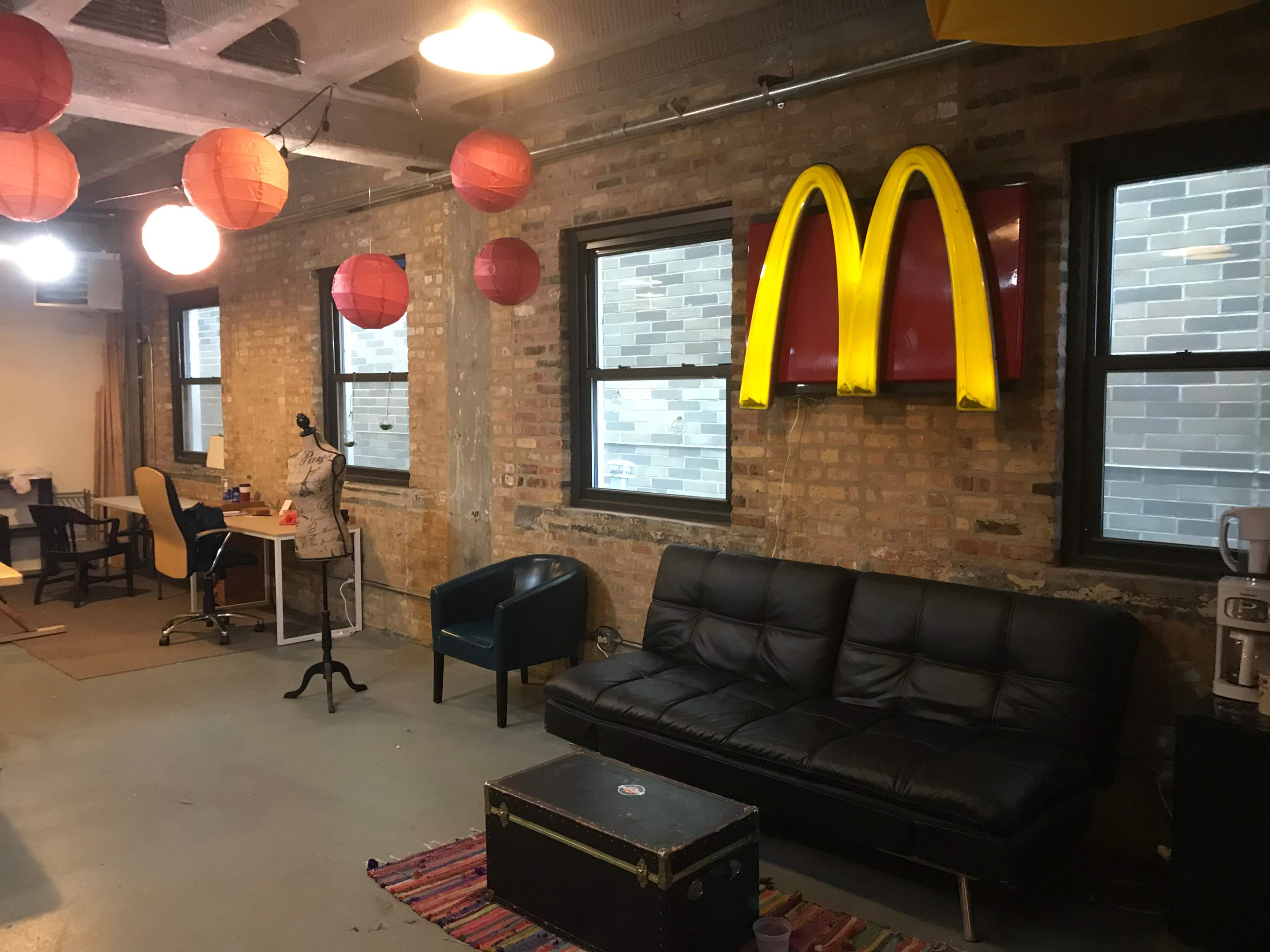 The image shows a casual office space featuring a black couch, a coffee table, a desk with a chair, and hanging red lanterns, with a large McDonald's logo displayed on the wall.