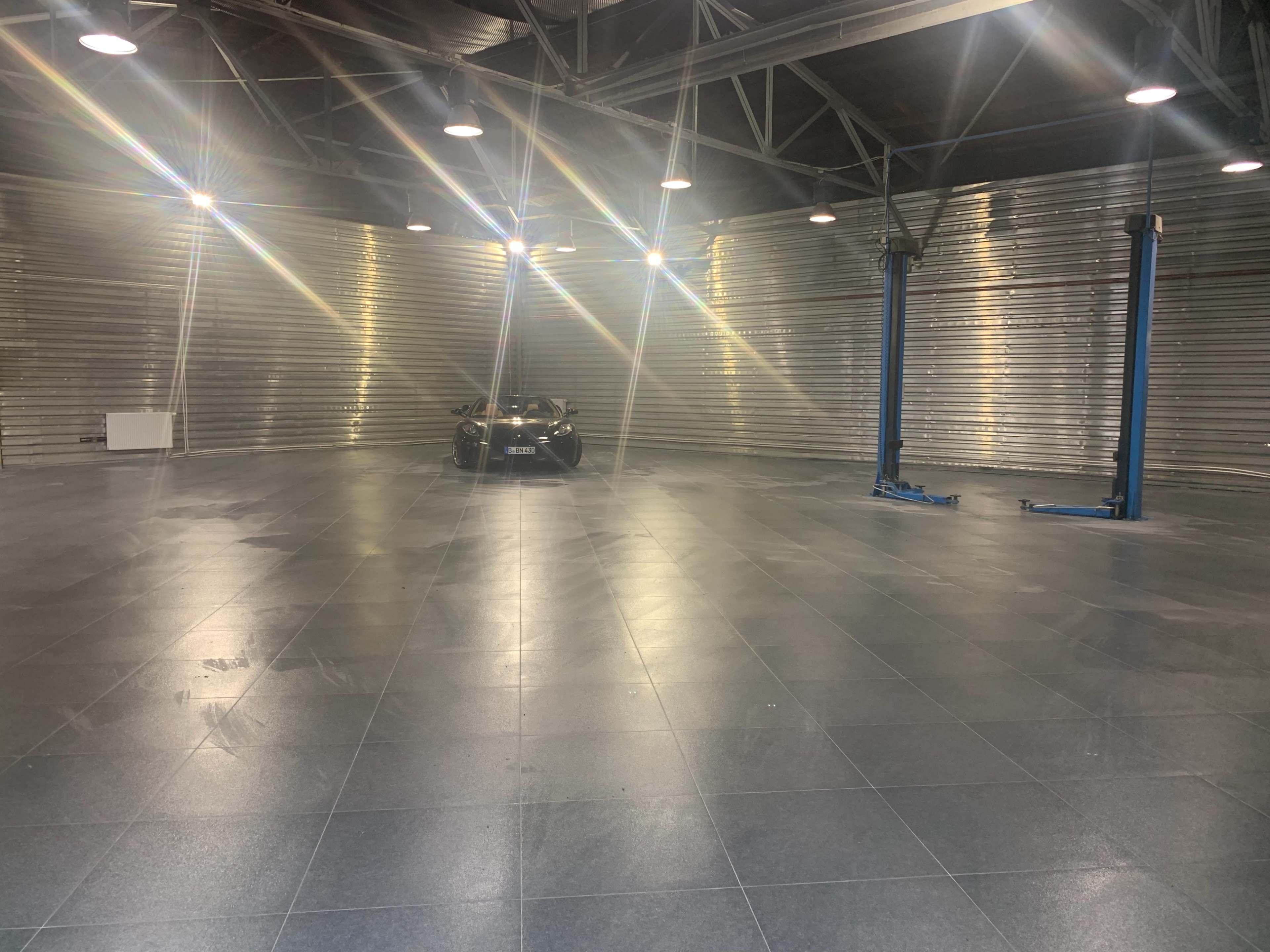 A black car is parked in the center of a spacious, well-lit garage with metallic walls and two vehicle lifts.