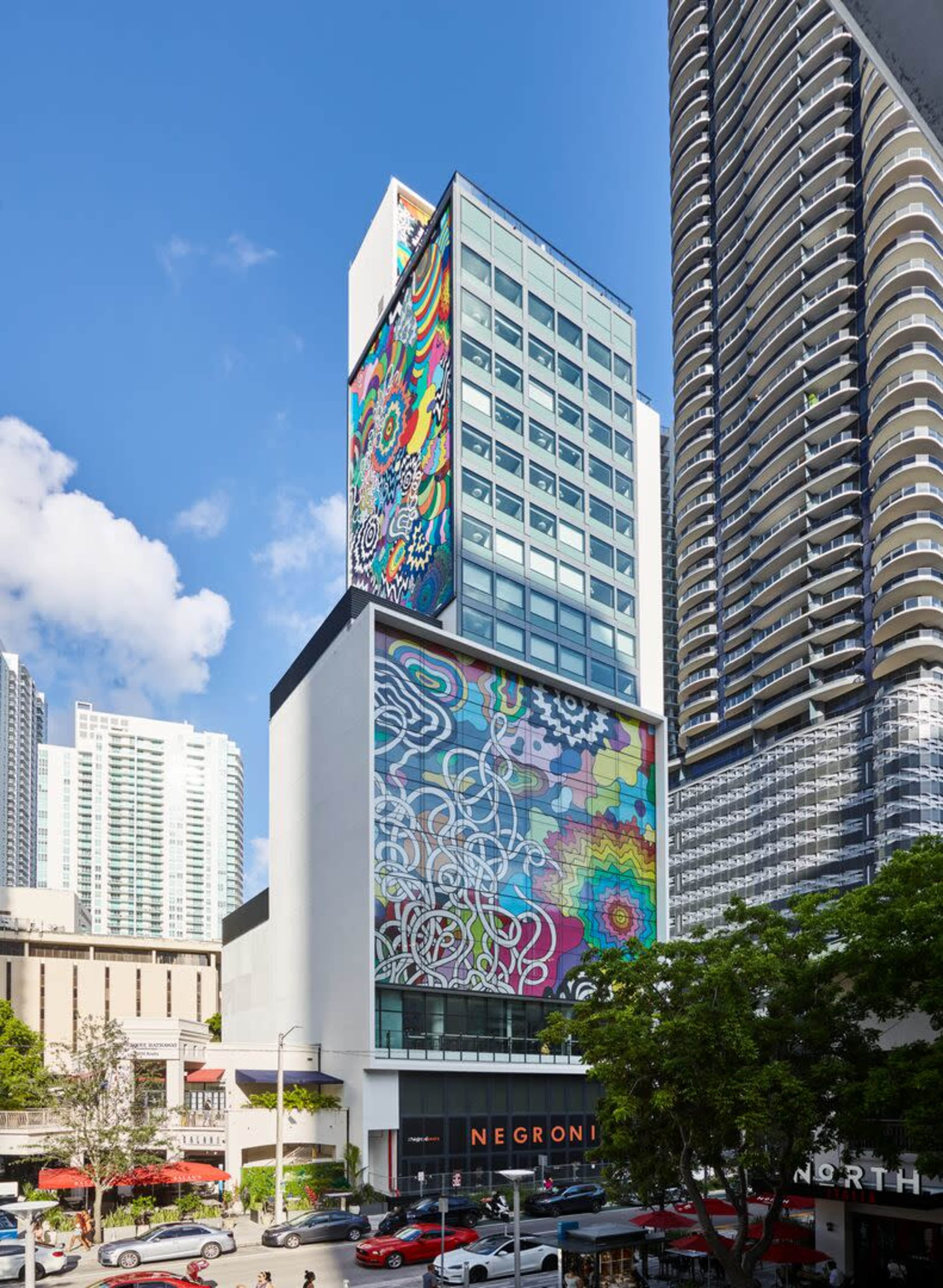 A modern building with a colorful mural on its exterior stands next to a high-rise structure in an urban setting.