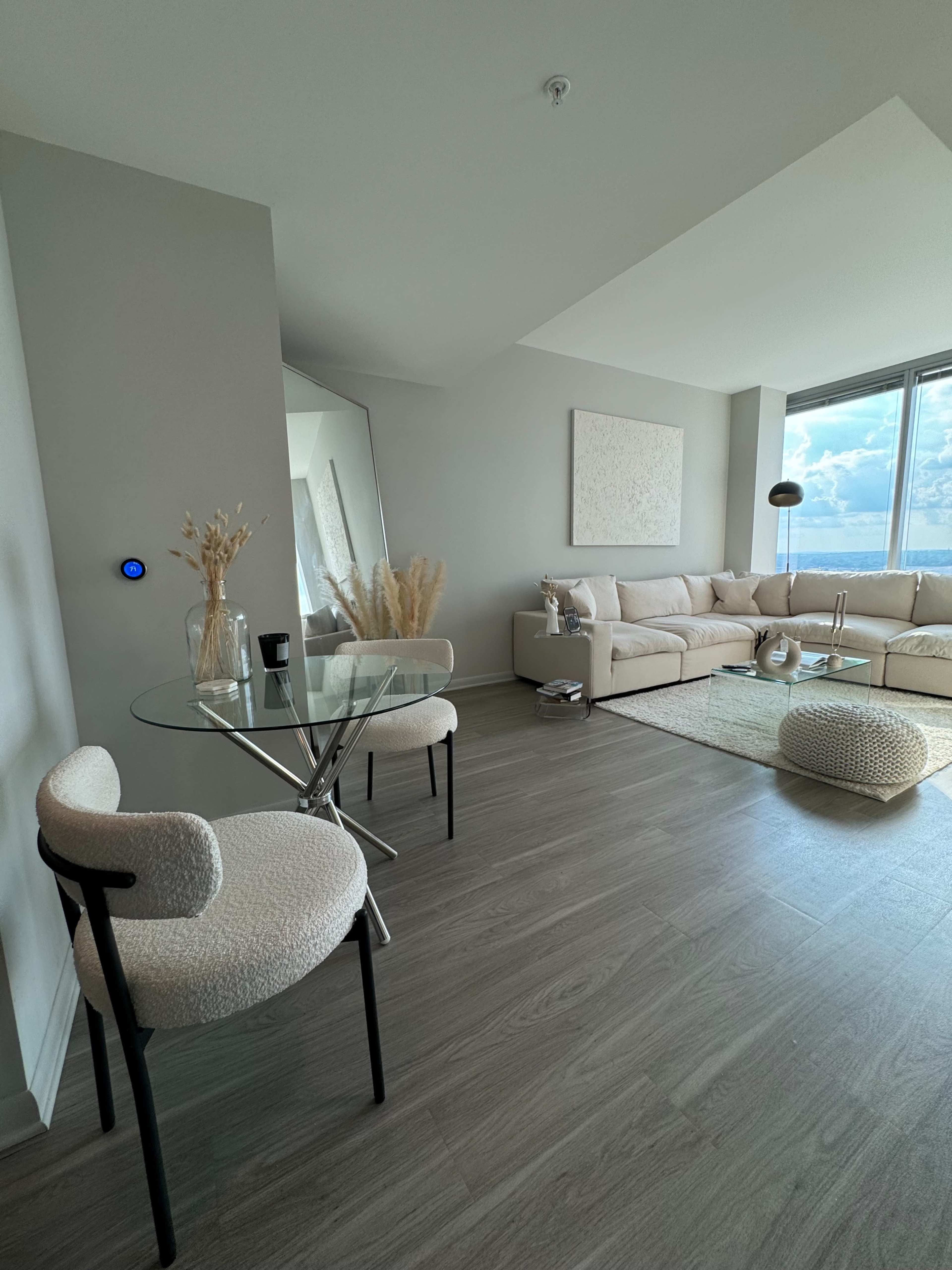 A modern living room features a beige sectional sofa, a glass coffee table, and two chairs, with large windows showcasing a view outside.