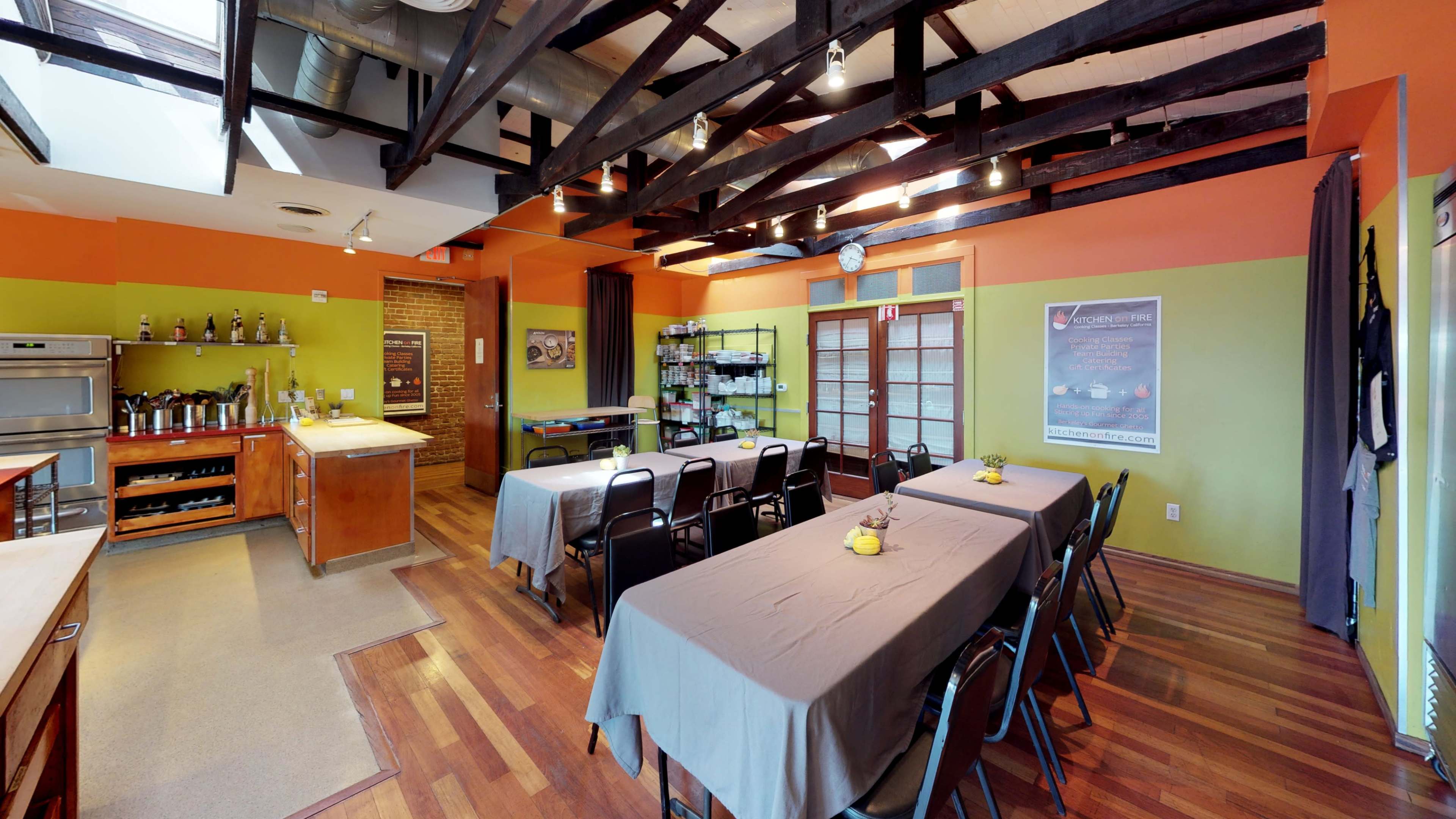 The image shows a food preparation area with several tables covered in gray tablecloths, surrounded by black chairs, and features wooden beams on the ceiling.