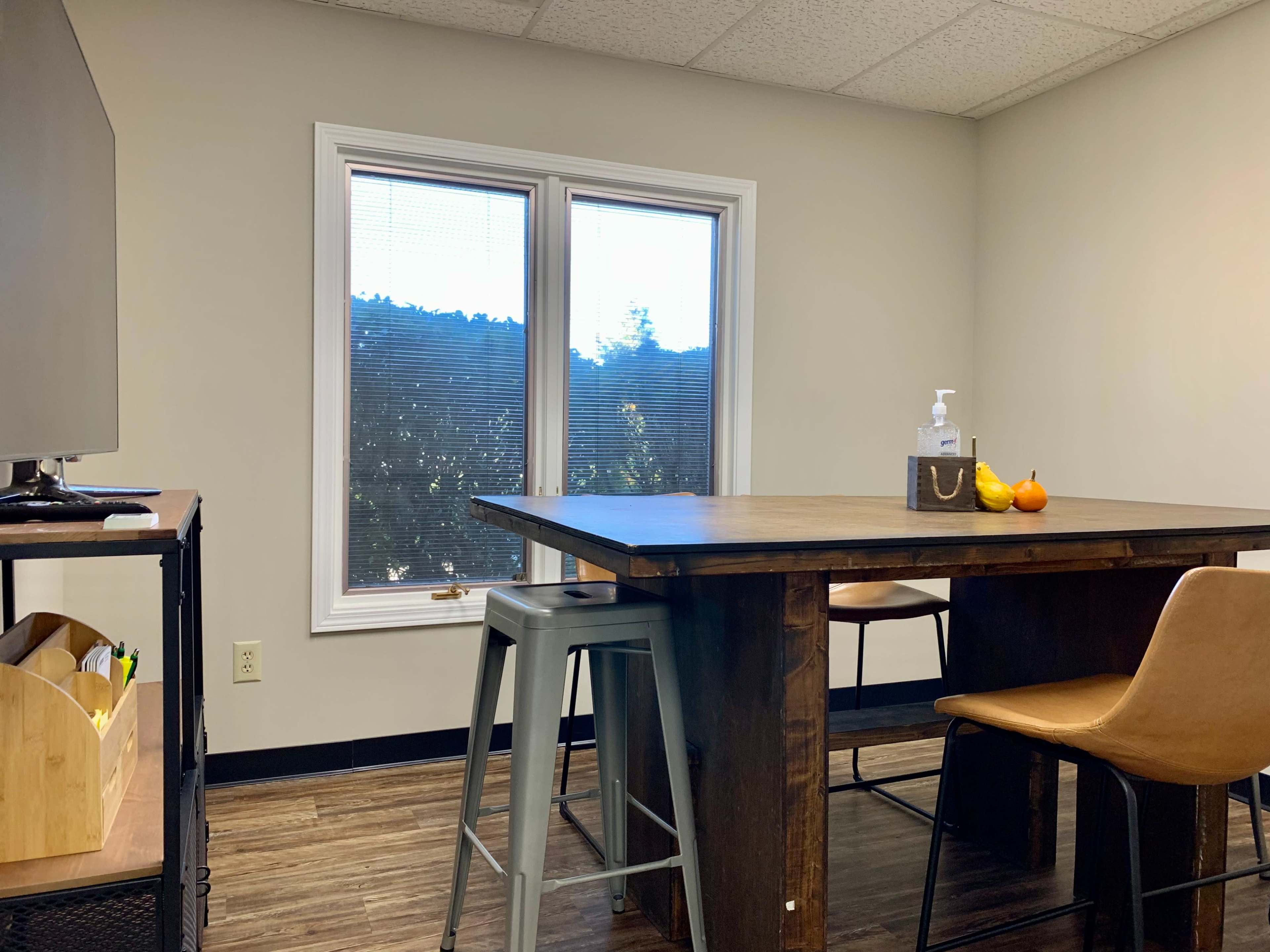 A wooden table with high stools sits in a well-lit room featuring a large window overlooking a wooded area.