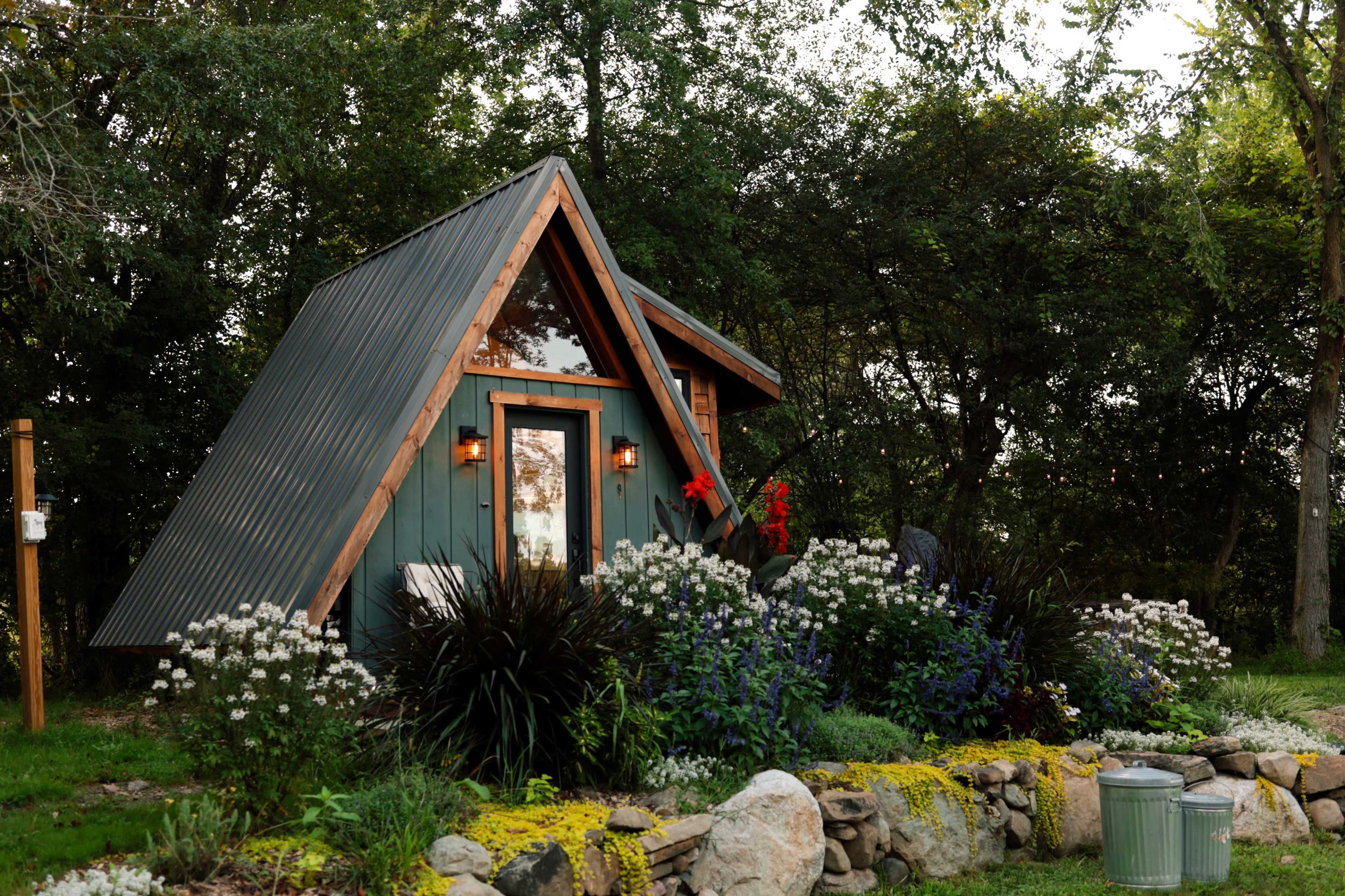 A small A-frame cabin is surrounded by a colorful flower garden and lush greenery.