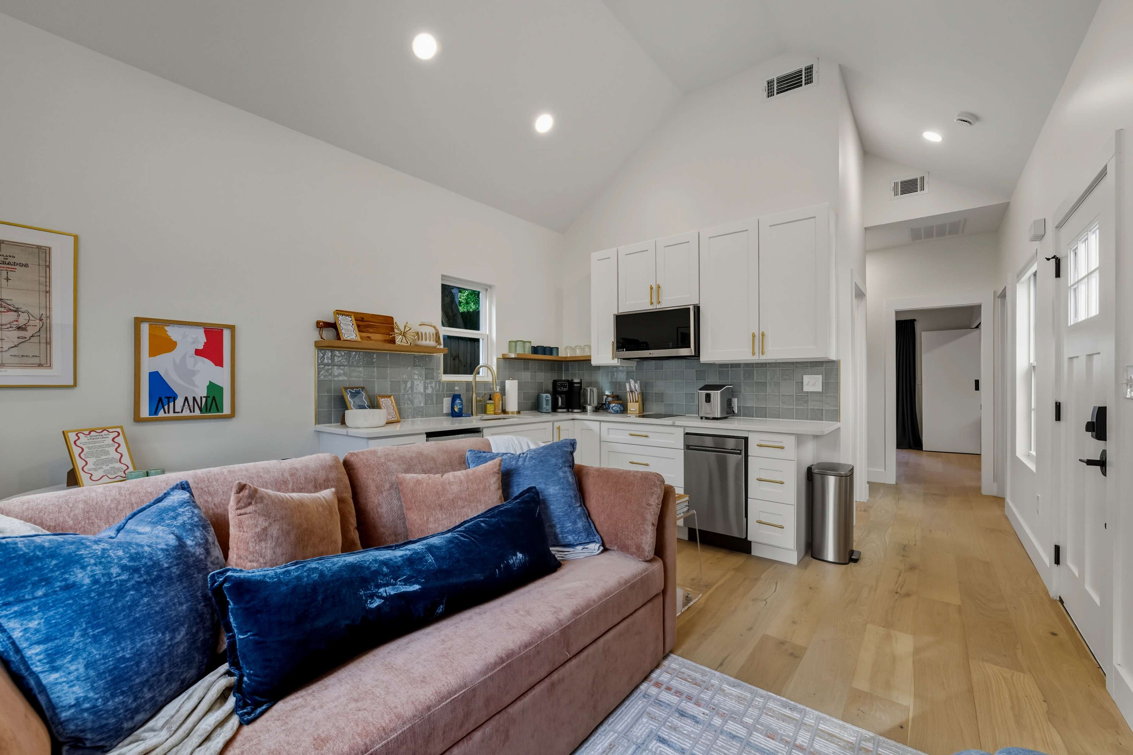 A modern living space features a pink sofa adorned with blue pillows, adjacent to a kitchenette with white cabinets and stainless steel appliances.