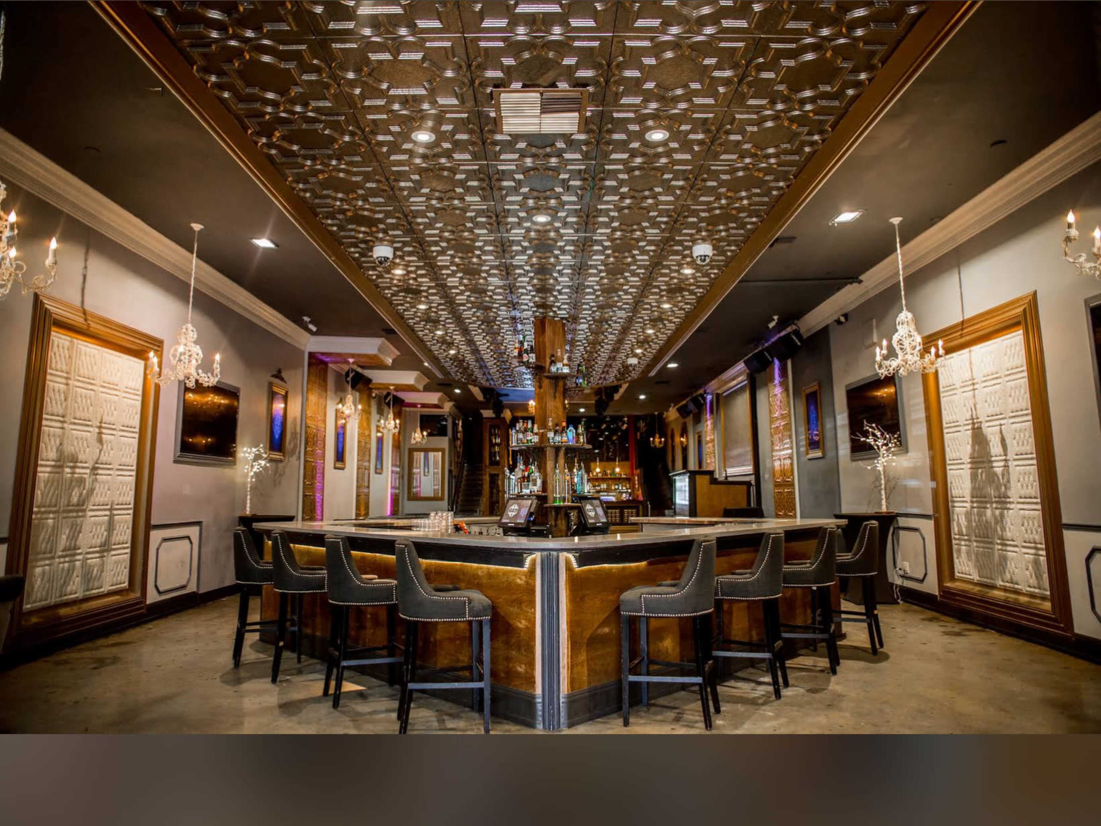 The image shows a spacious bar area with a central counter, elegant chandeliers, and decorative ceiling tiles.