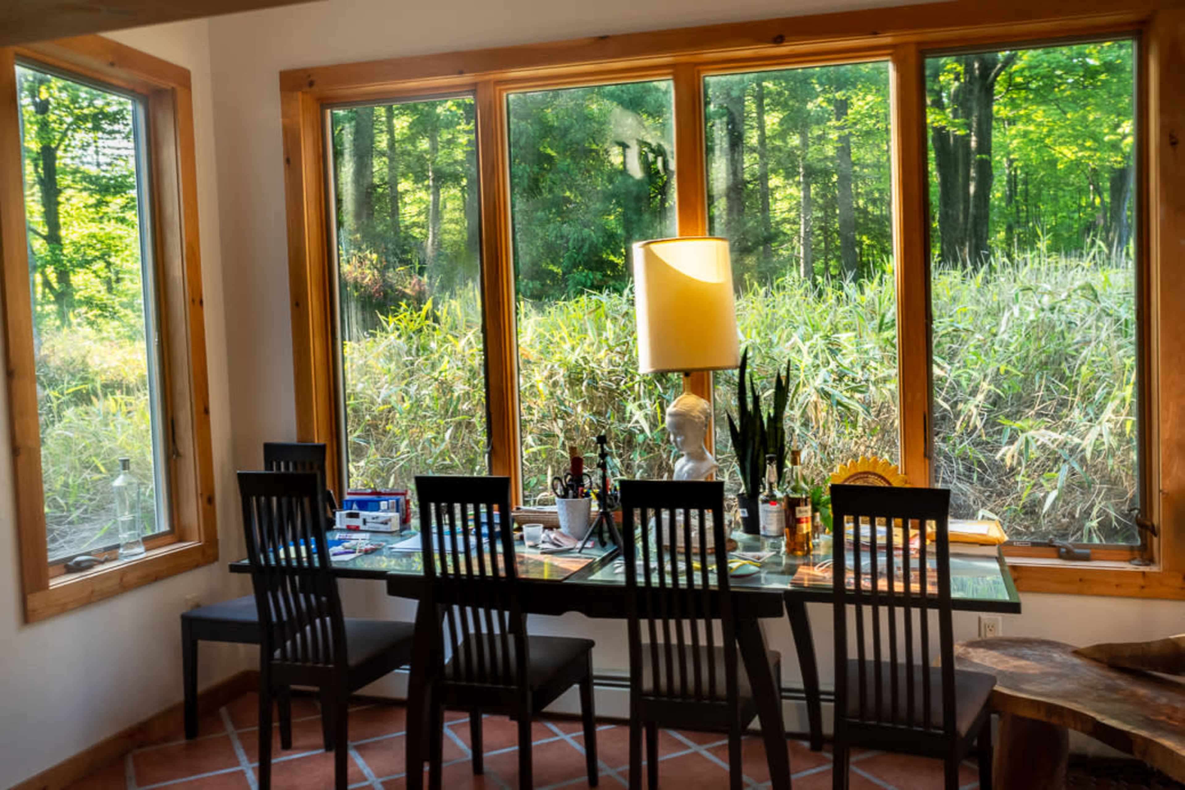 A dining table with black chairs is positioned beside large windows that overlook a green, wooded area.
