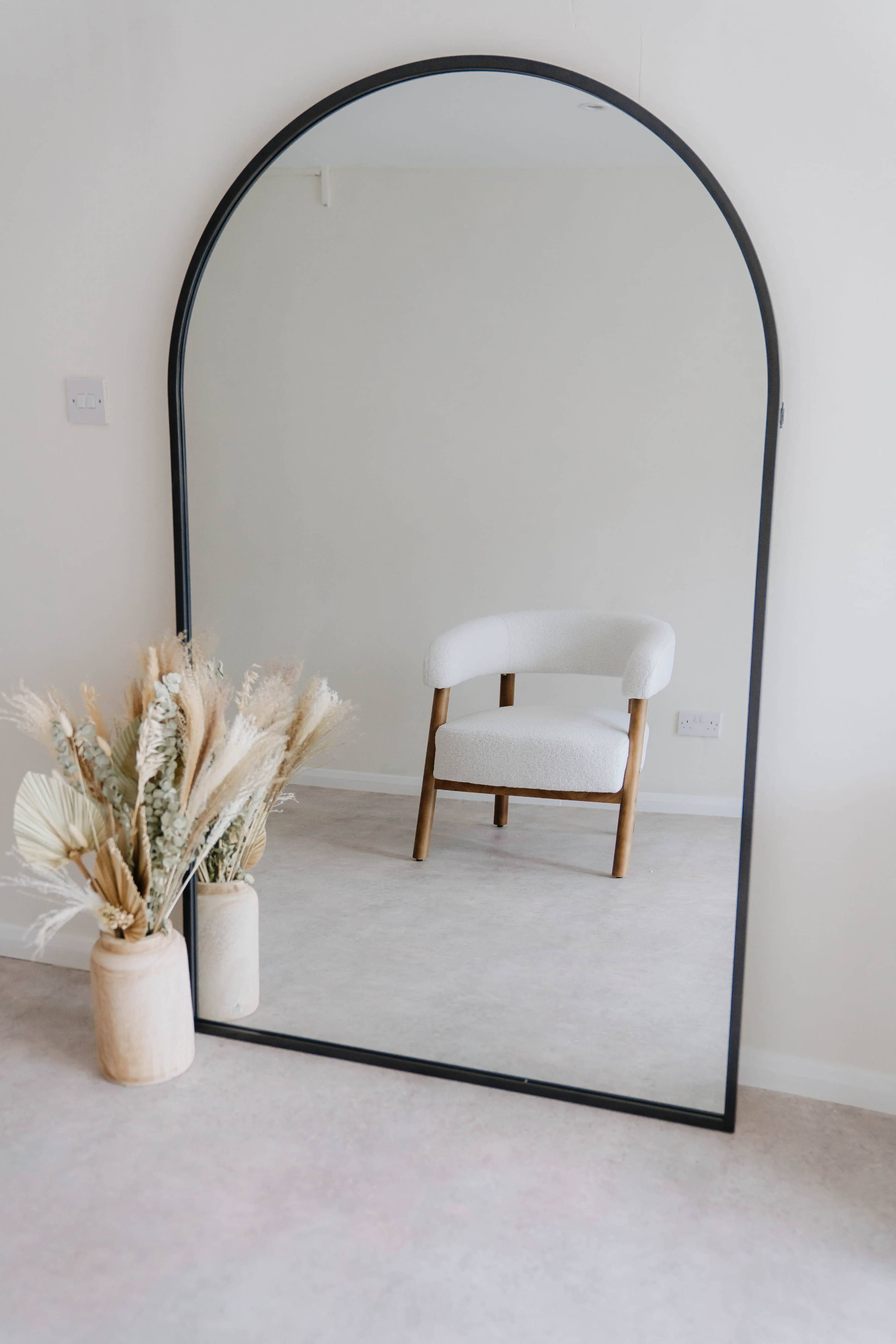 A large, arched mirror reflects a simple chair and a vase of dried flowers in a softly lit room.