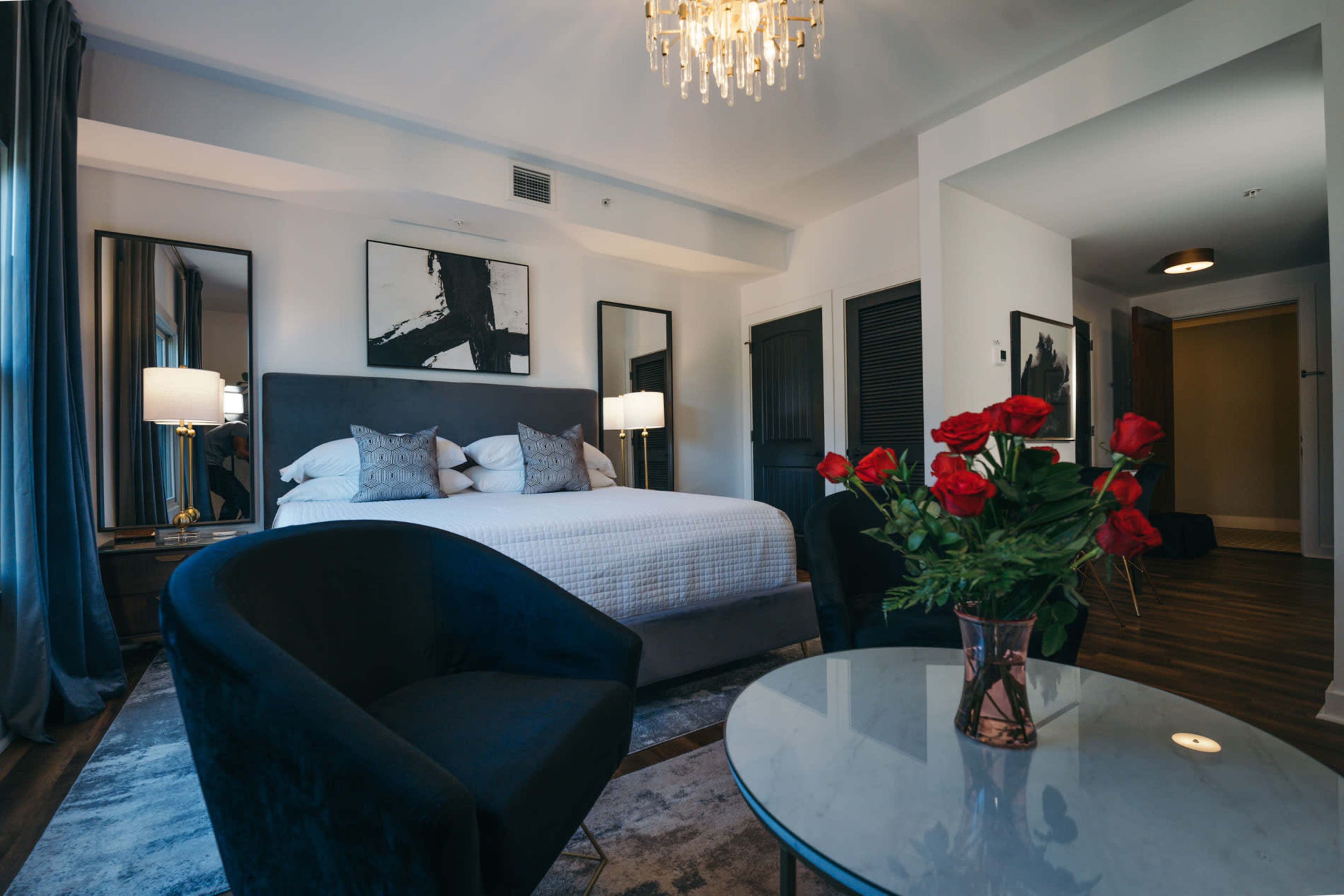 A modern hotel room features a king-sized bed, two lamps, a seating area with a blue armchair, a glass table, and a bouquet of red roses.