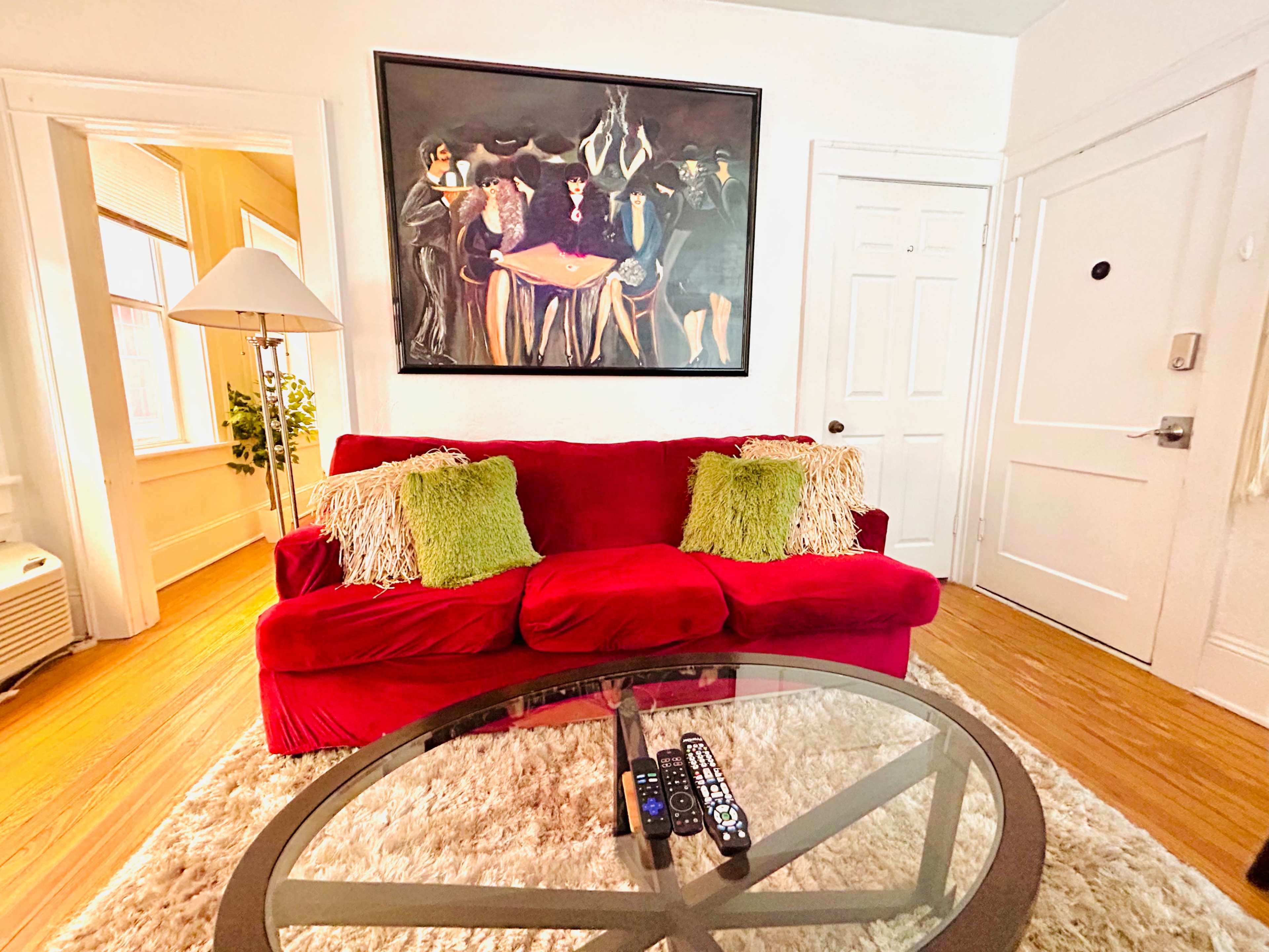 A living room featuring a red sofa with decorative green pillows, a glass coffee table with remote controls, and a large framed artwork on the wall.