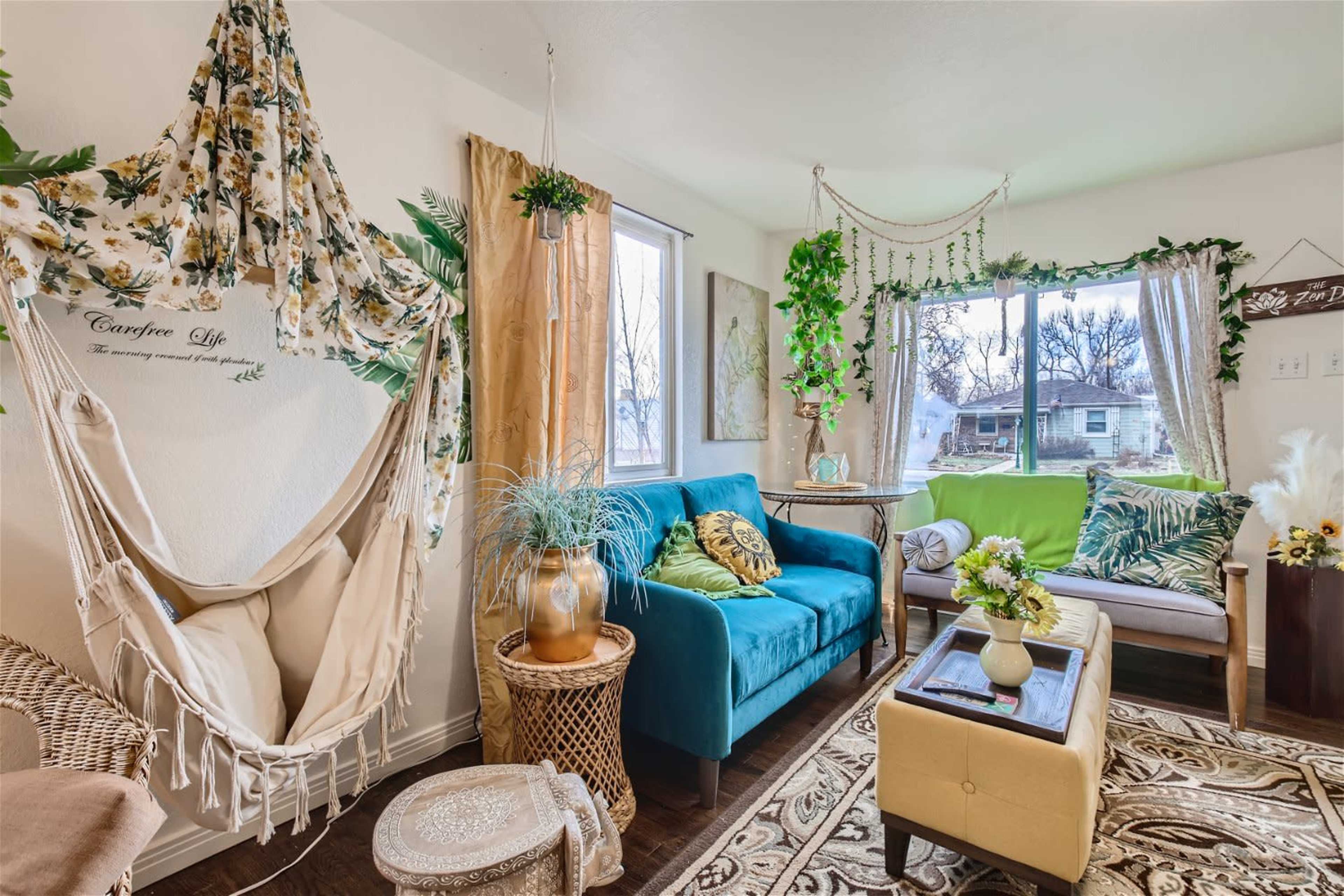 A cozy living room features a blue sofa, a hanging chair adorned with floral fabric, and large windows, with greenery and decorative elements throughout.