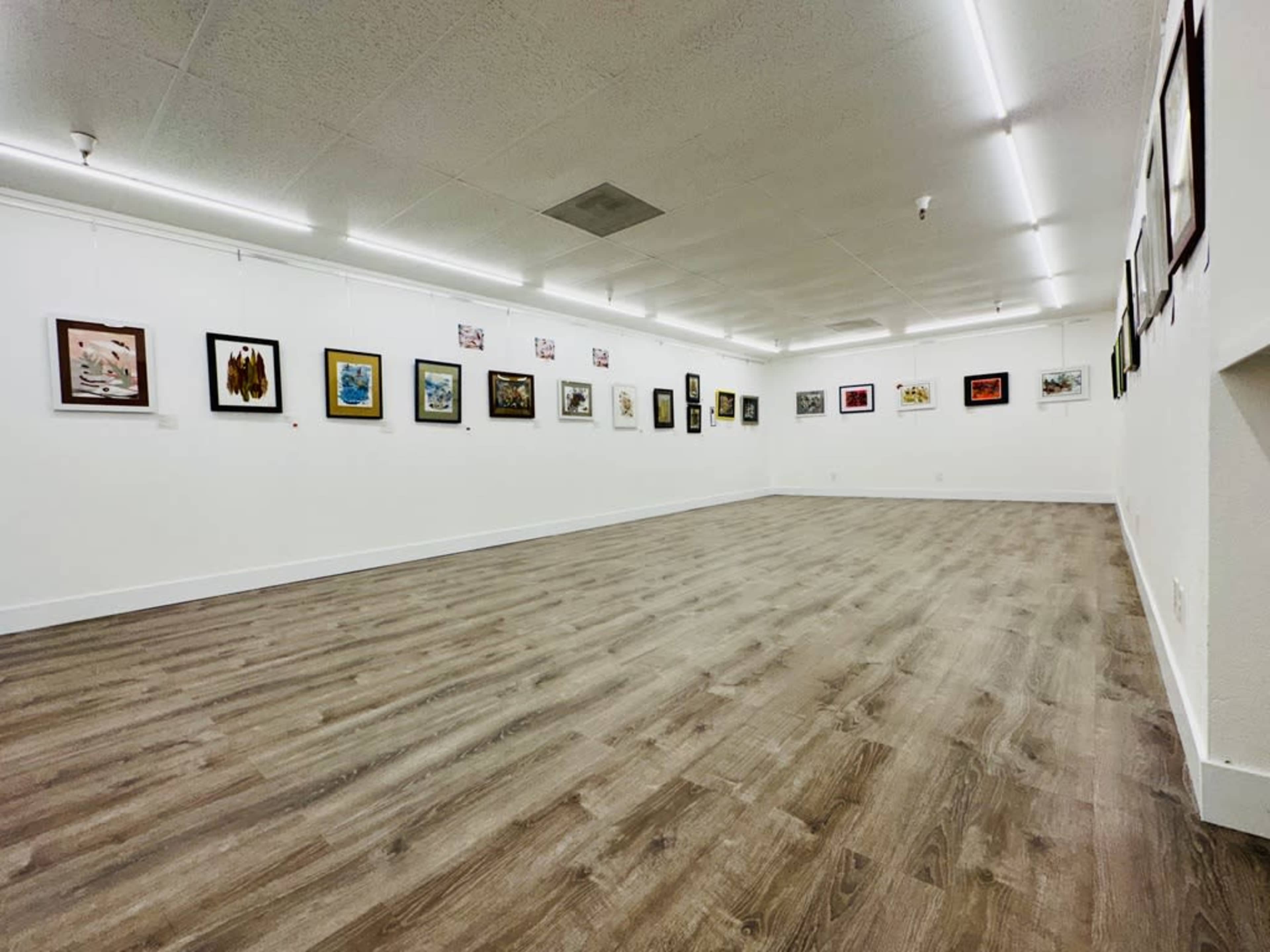 An art gallery with various framed artworks displayed on the white walls and a spacious wooden floor.