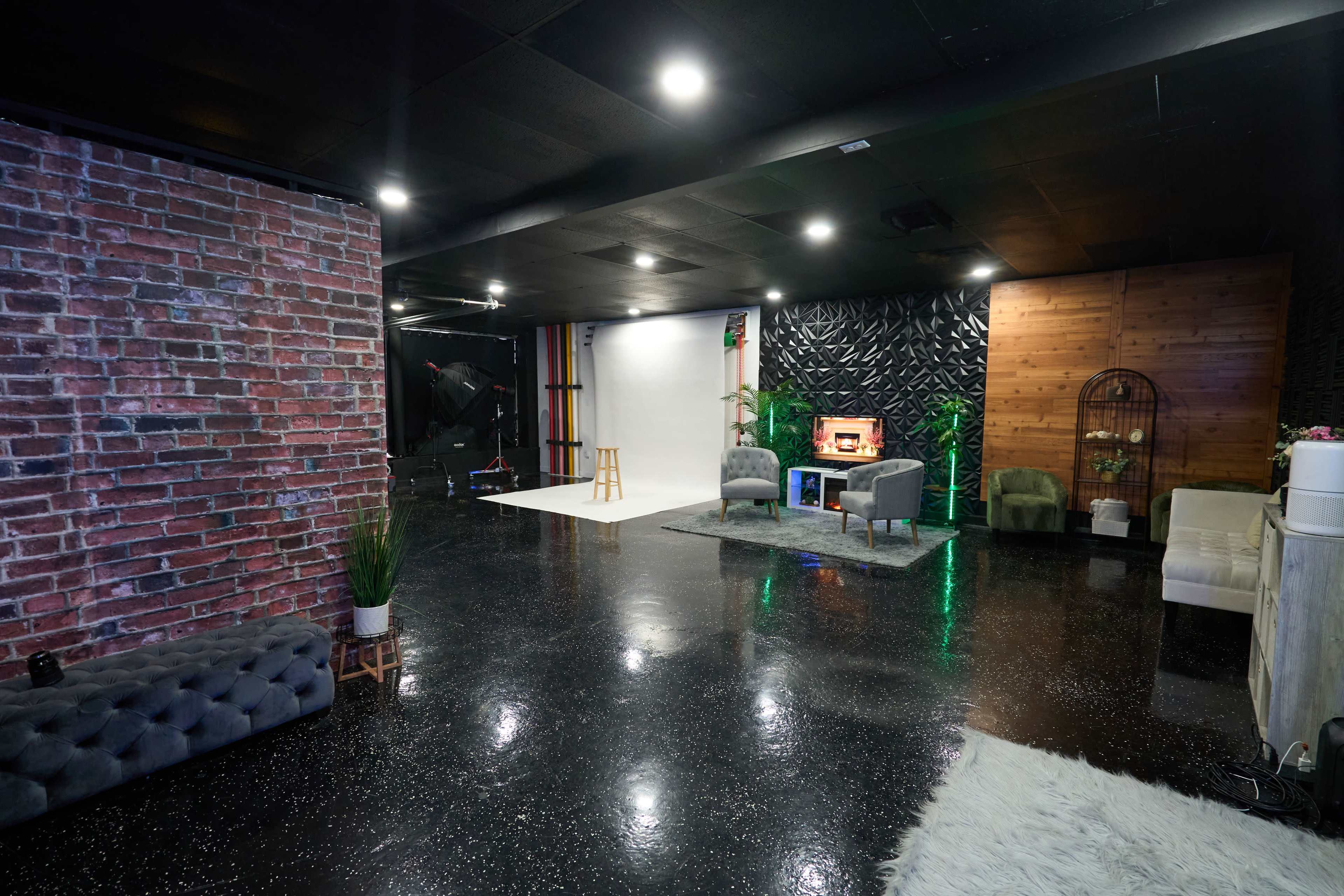 The image shows a modern studio space featuring a mix of brick, wood, and black textured walls, with various seating areas and a backdrop for photography or video production.