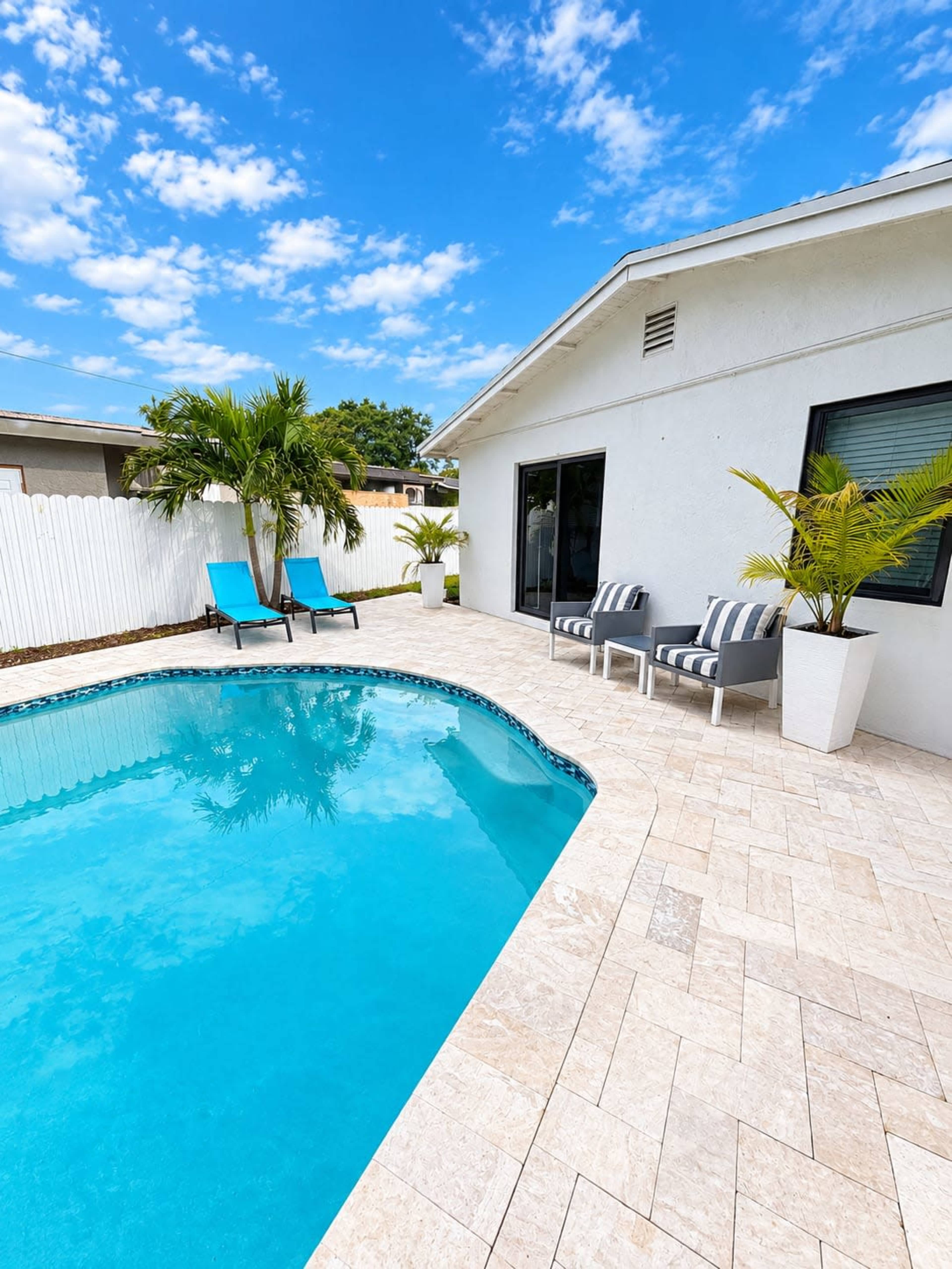 A clear blue pool is surrounded by stone tiles, lounge chairs, and potted plants beside a white house under a bright sky with scattered clouds.