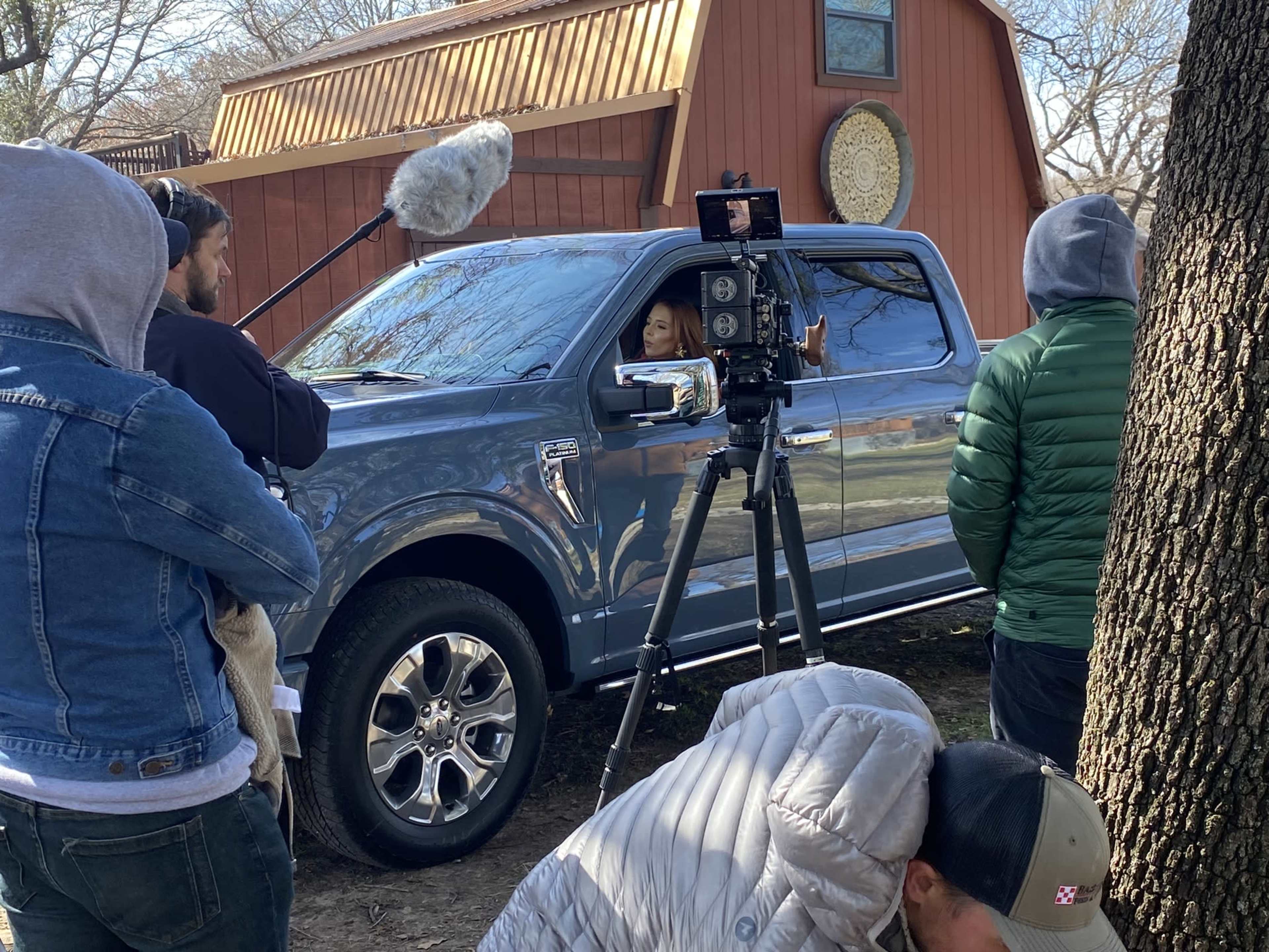 A film crew sets up equipment near a gray pickup truck parked in front of a barn.