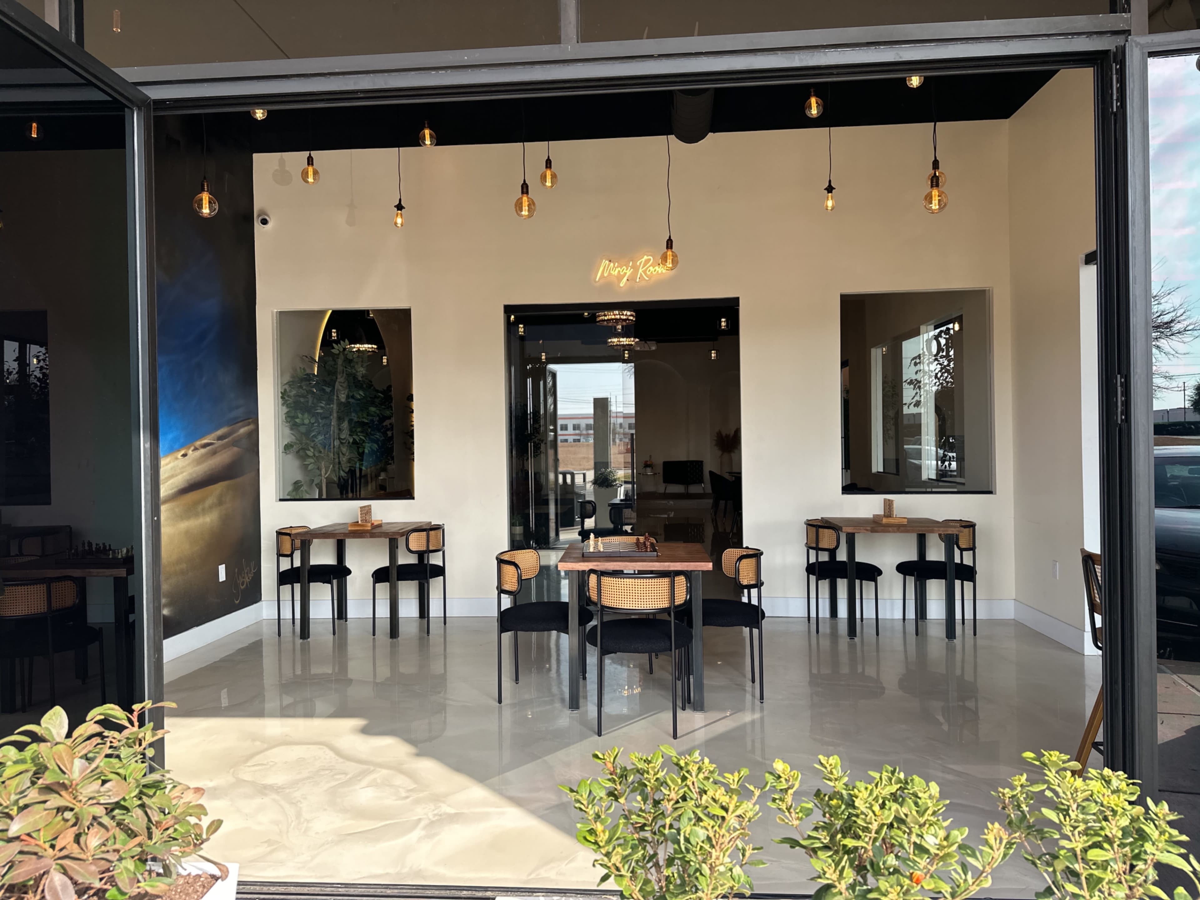 The image shows the interior of a restaurant with several tables and chairs arranged neatly, along with decorative lighting elements hanging from the ceiling.