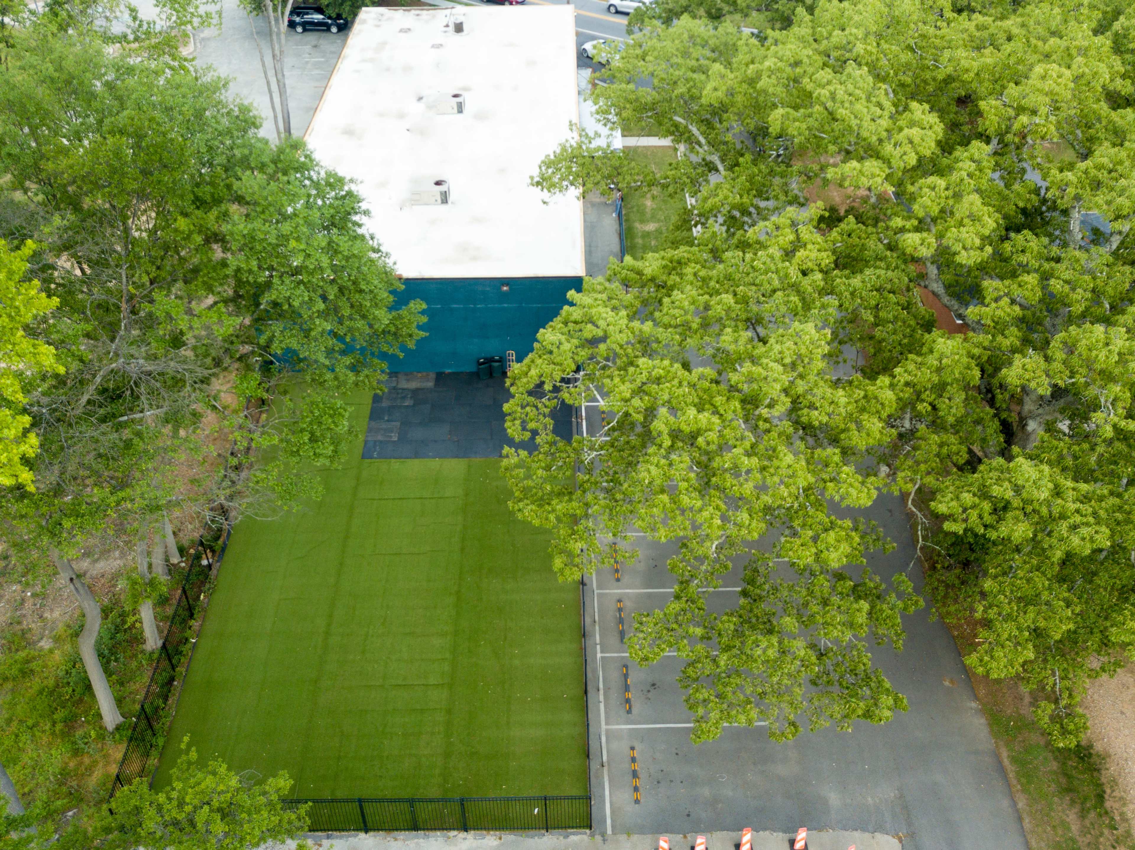 An aerial view shows a green artificial turf area adjacent to a building surrounded by trees and a paved parking lot.