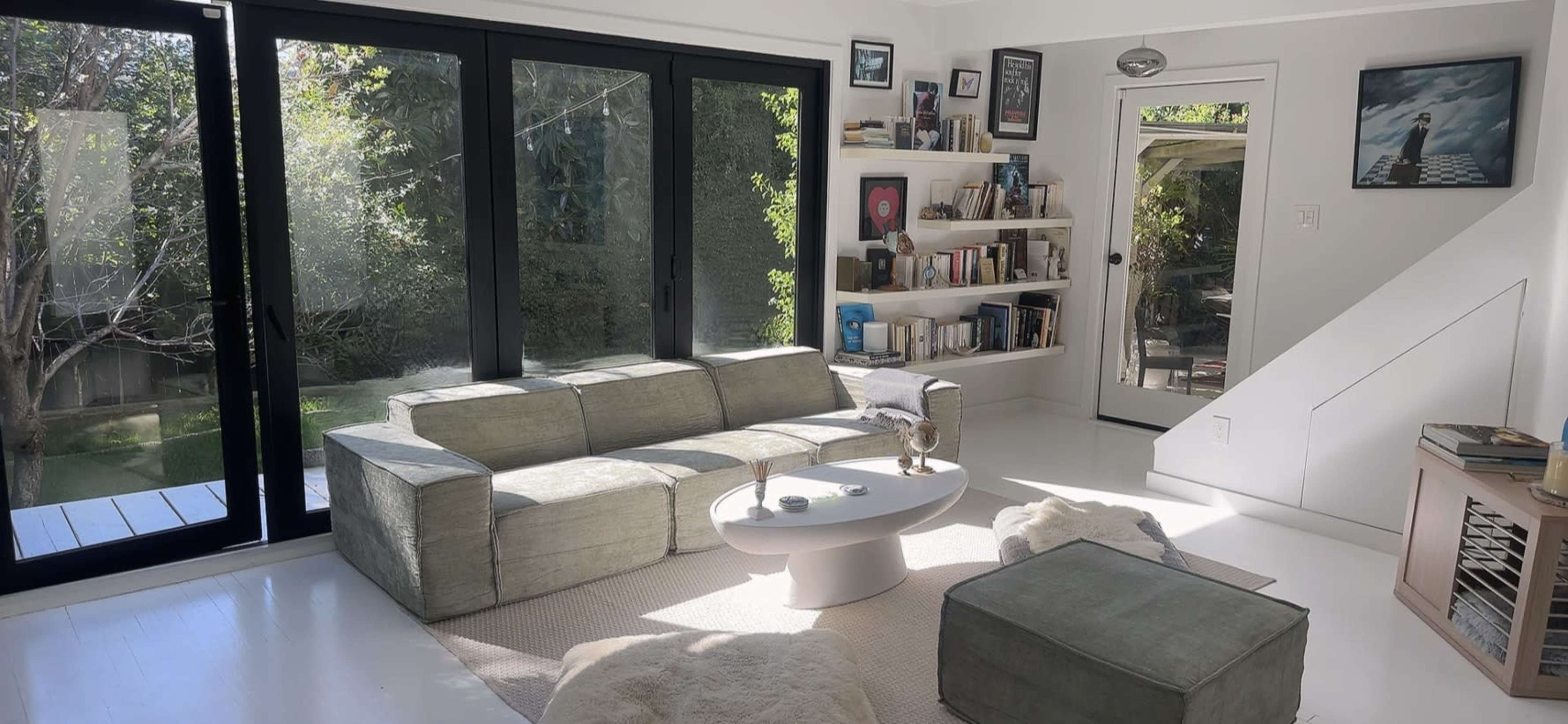 The image shows a modern living room with large windows, a light-colored couch, a round coffee table, and a bookshelf, all set against a backdrop of greenery outside.