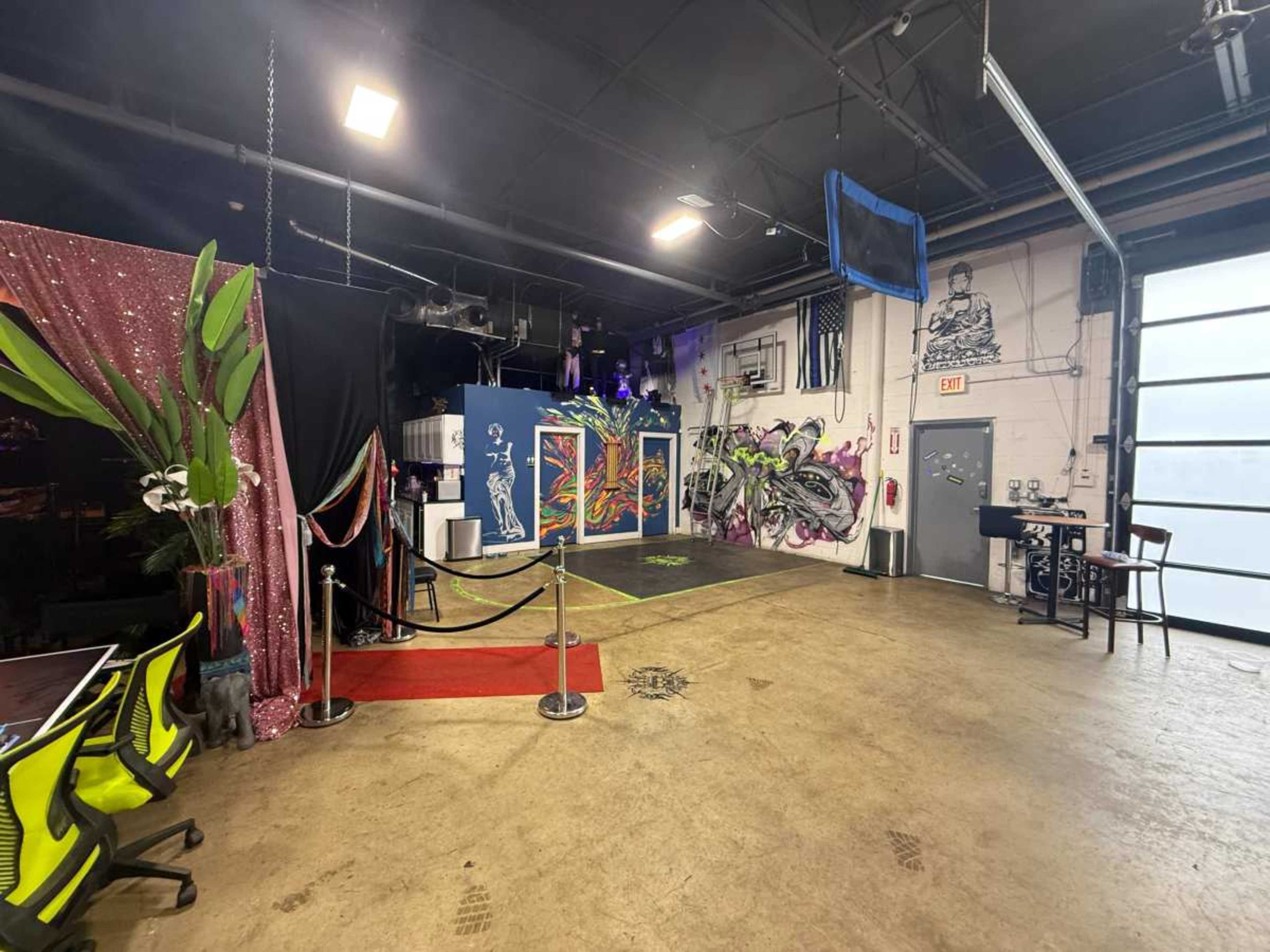 The image shows a spacious indoor venue featuring a colorful mural on one wall, a red carpet leading to a small stage area, and decorative elements including plants and string lights.