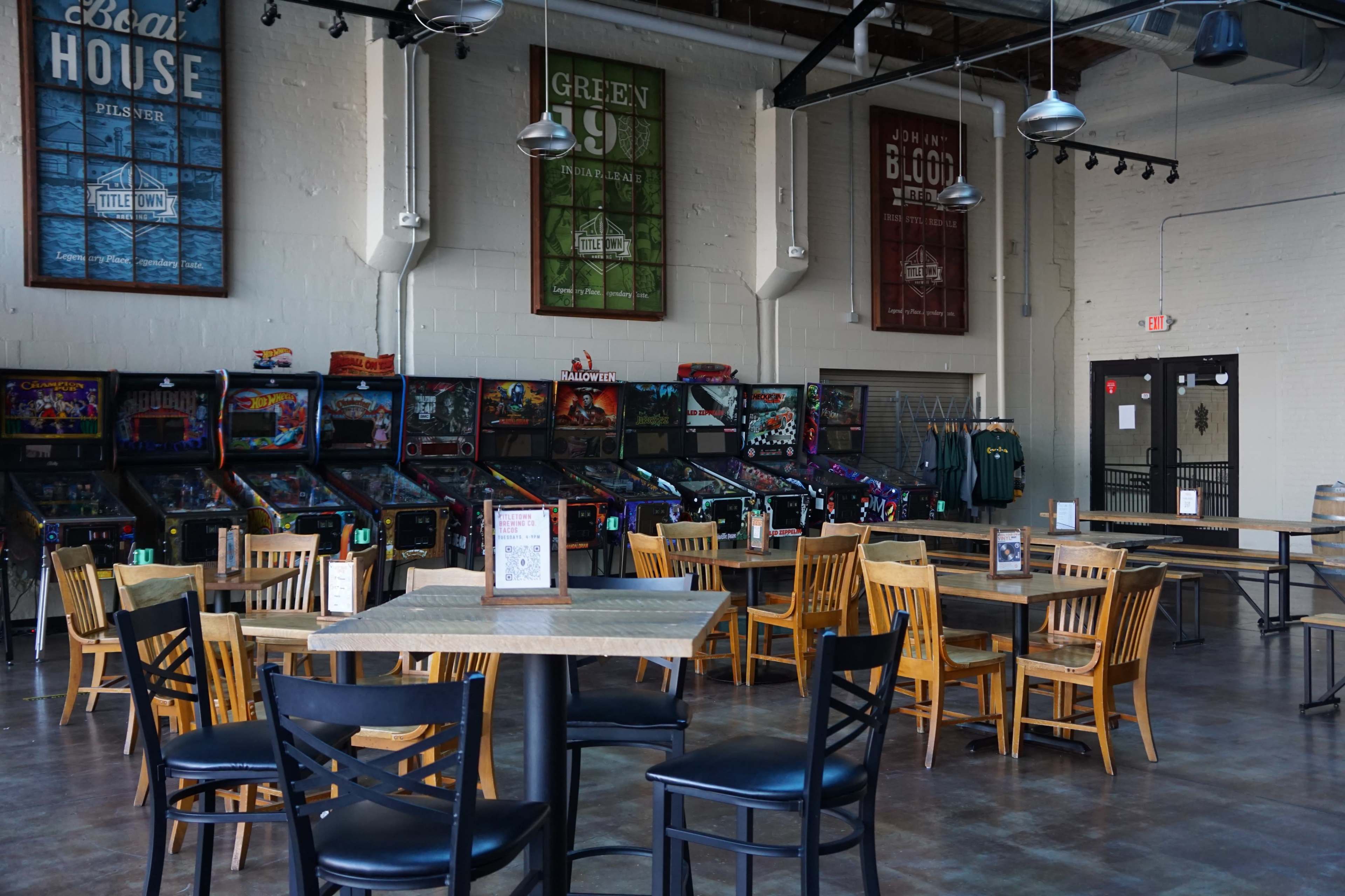 A spacious game room with several pinball machines lined against the wall and wooden tables and chairs arranged throughout the area.