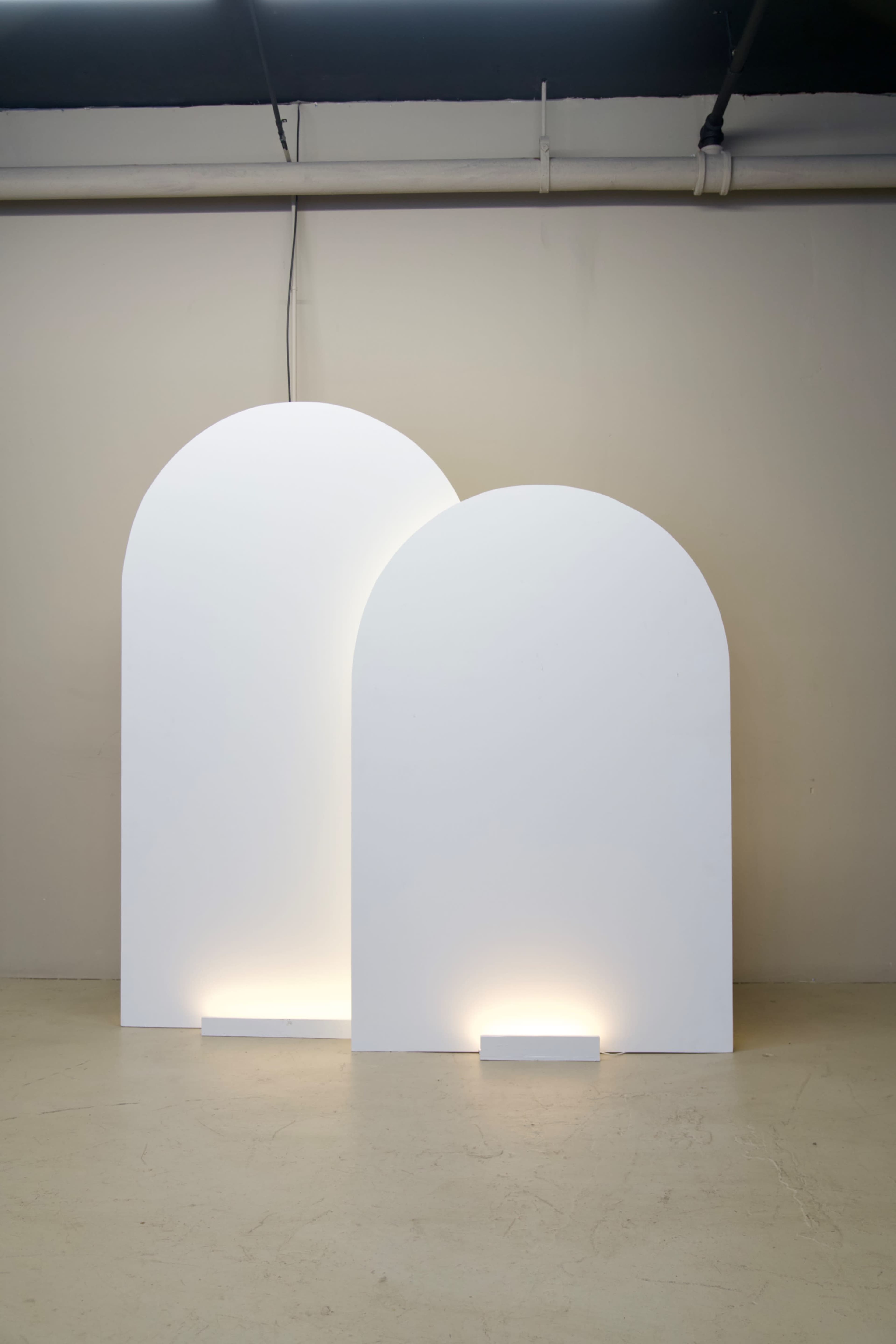 Two large, white, arch-shaped panels are illuminated from below against a neutral-colored wall.