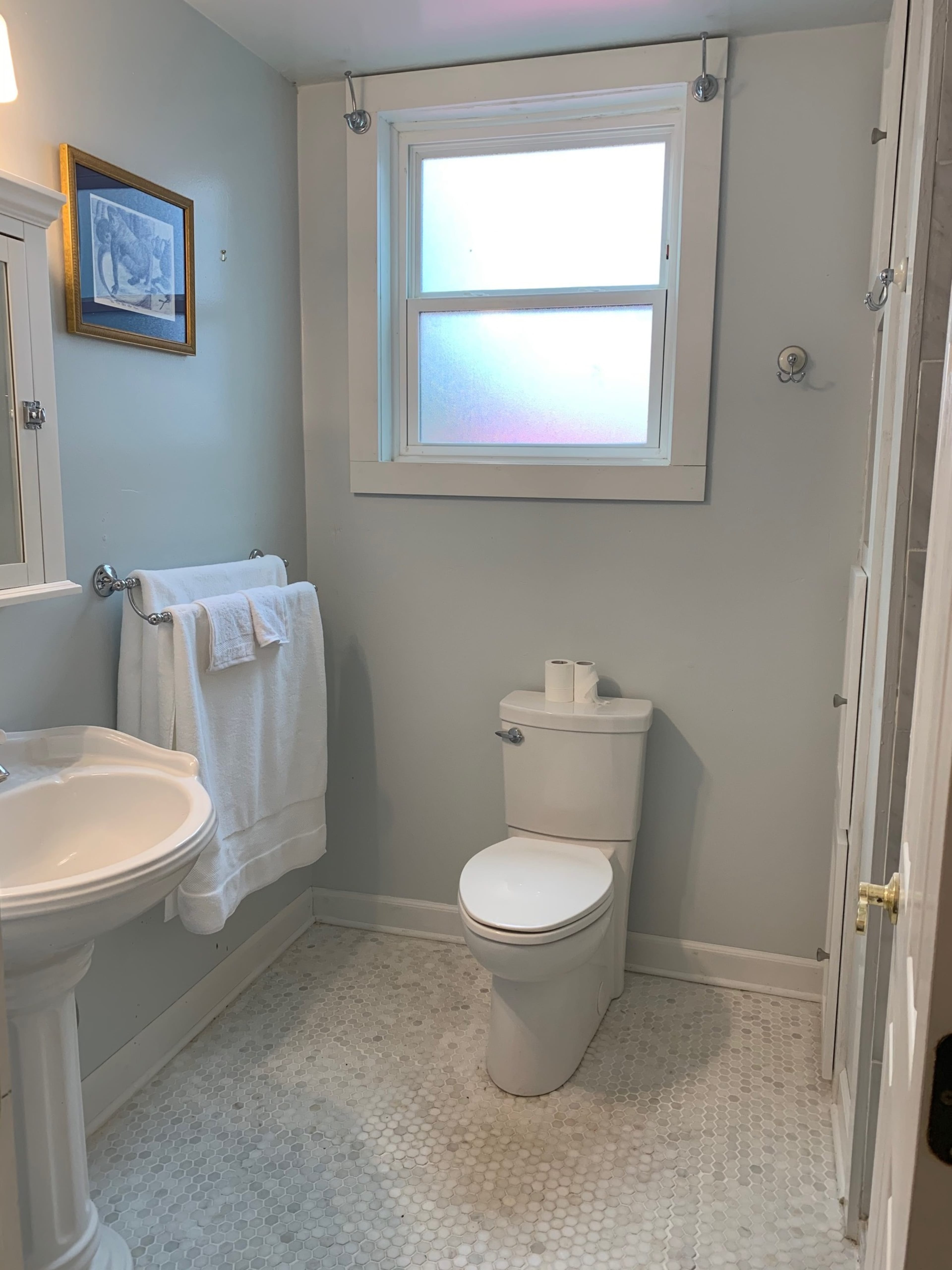 The image shows a small bathroom with a pedestal sink, a toilet, a window, and white towels displayed on a rack.