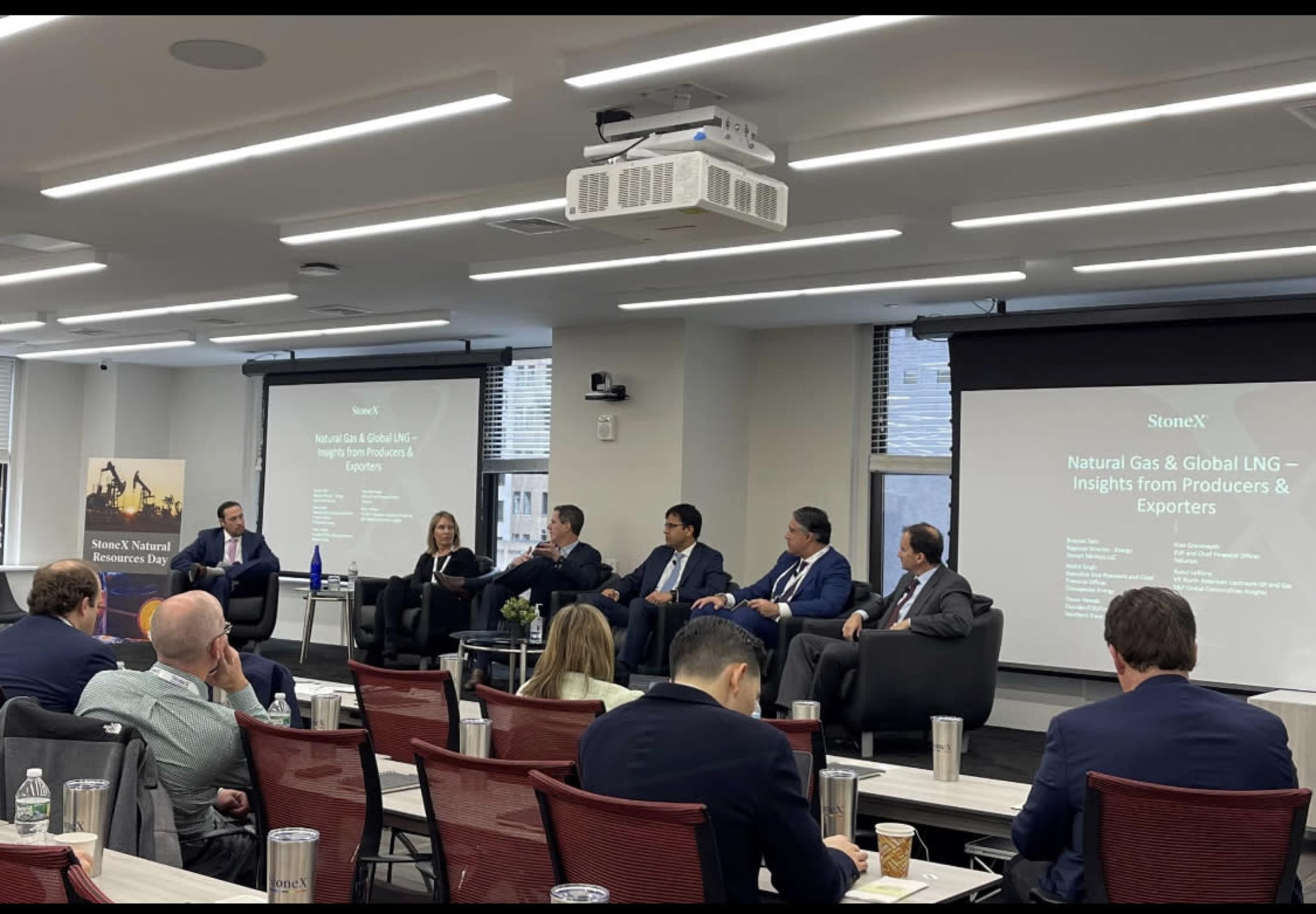 A panel discussion is taking place in a conference room, featuring six speakers seated on a stage with a presentation displayed on the screen behind them.