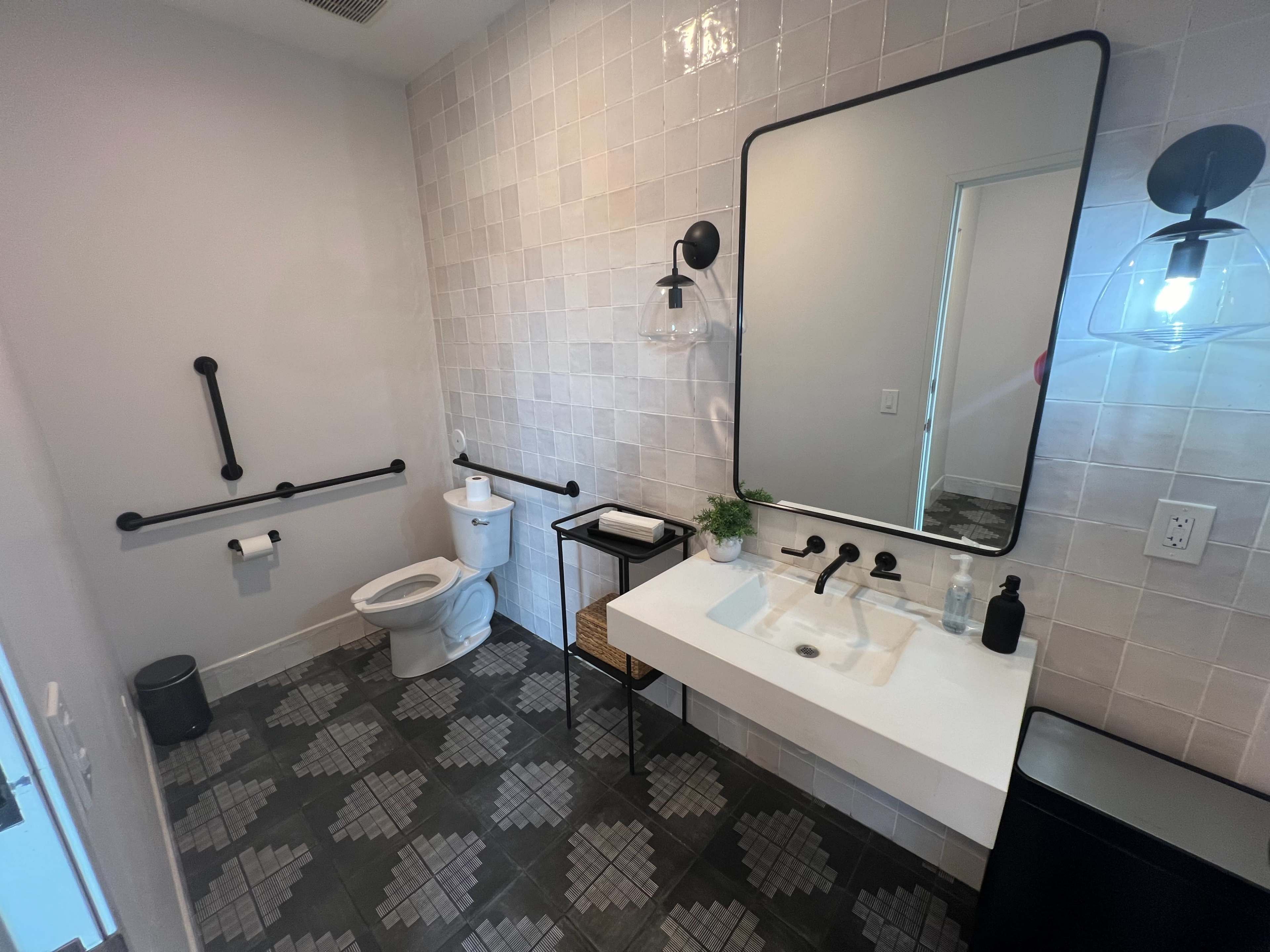 A modern bathroom features a toilet, a rectangular sink with a mirror above it, and patterned tile flooring, along with black grab bars and lighting fixtures.