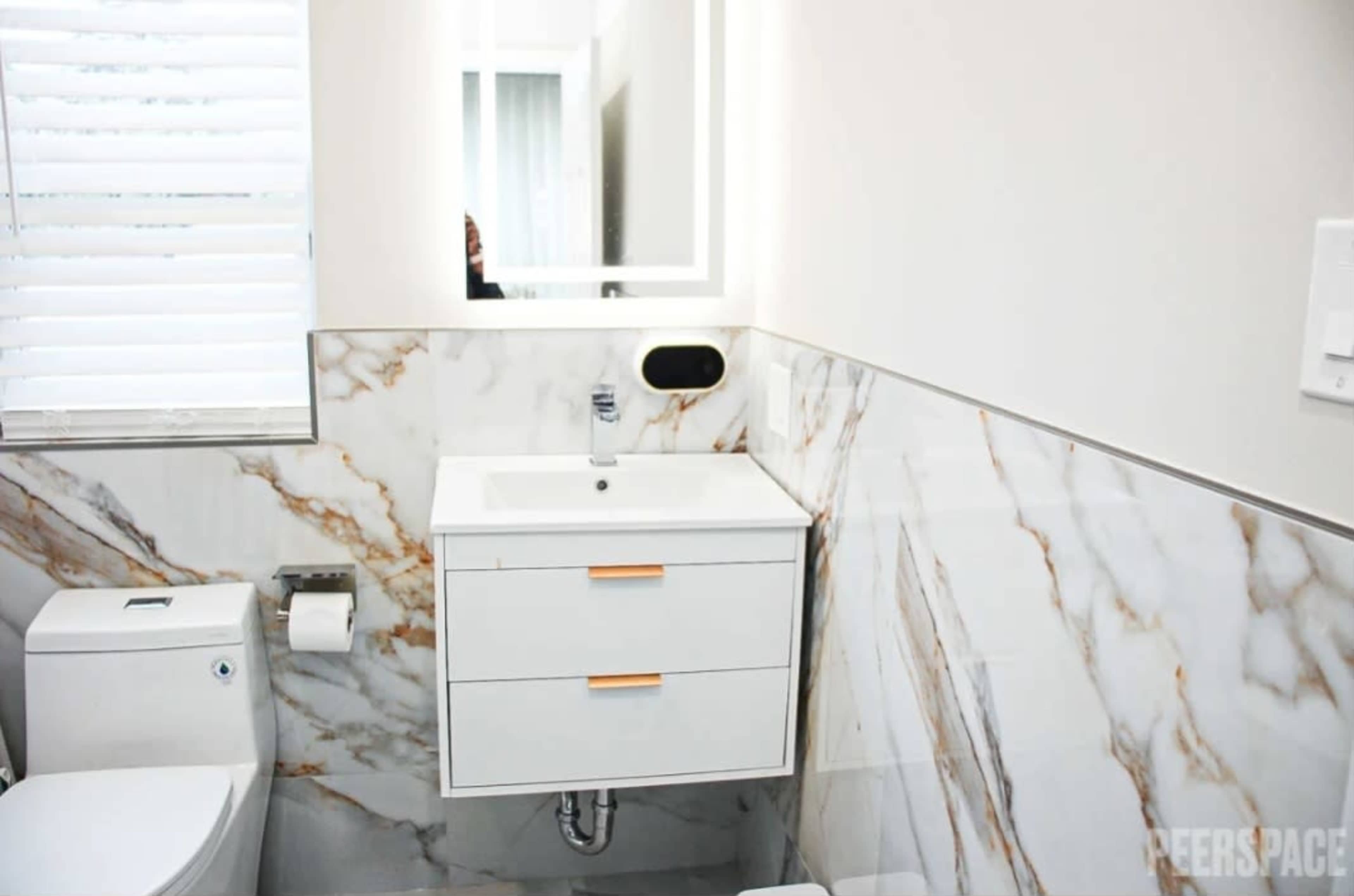 A modern bathroom features a white vanity with two drawers, a mirror with integrated lighting, and marble-patterned walls.