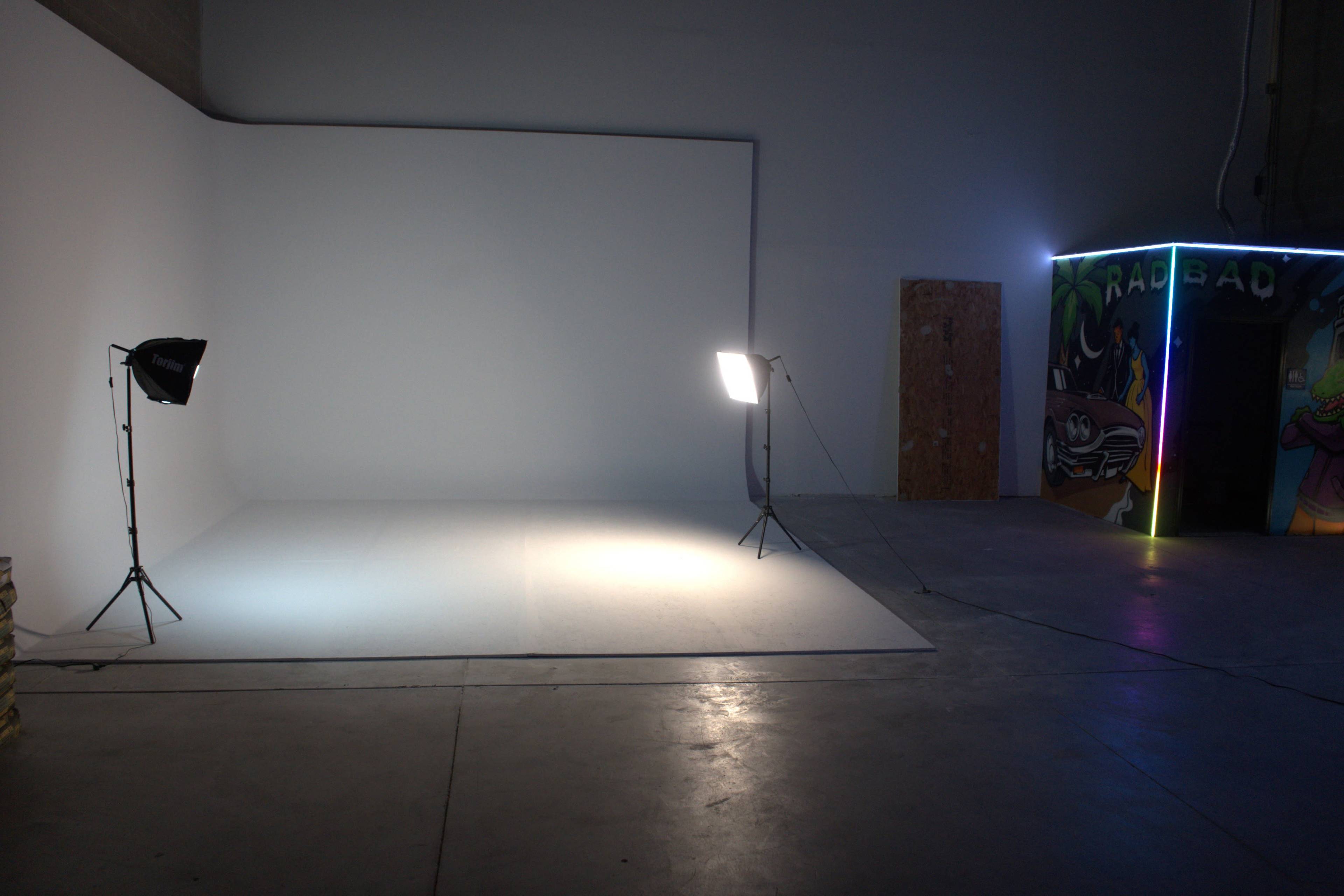 The image shows a minimalistic studio space with a white backdrop, two lighting setups, and a colorful corner featuring graffiti art.