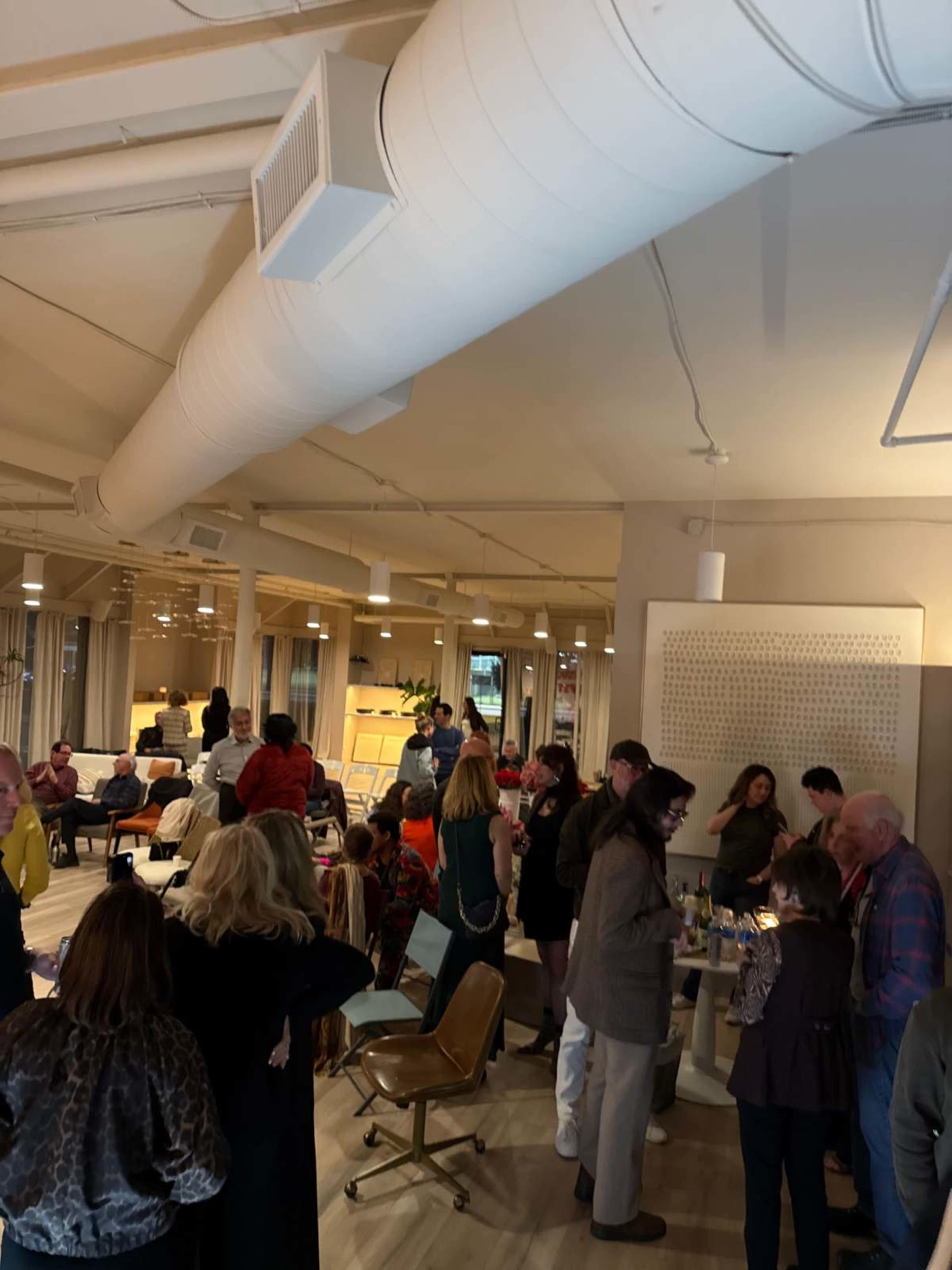 A diverse group of people gathers in a modern, well-lit workspace, engaging in conversation around tables and seating areas.