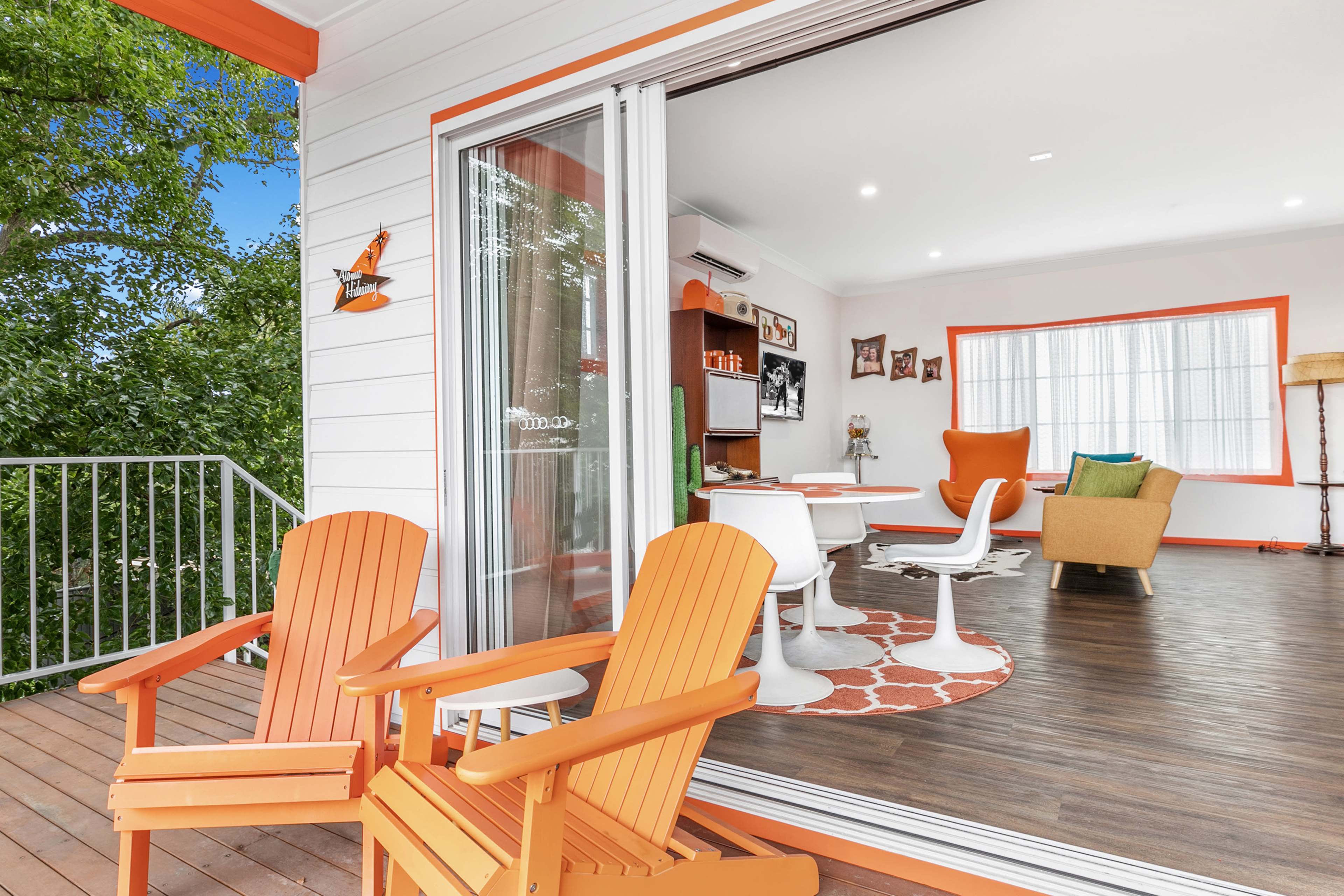 An outdoor balcony with two orange chairs, leading into a brightly lit living room featuring white walls and colorful furniture.