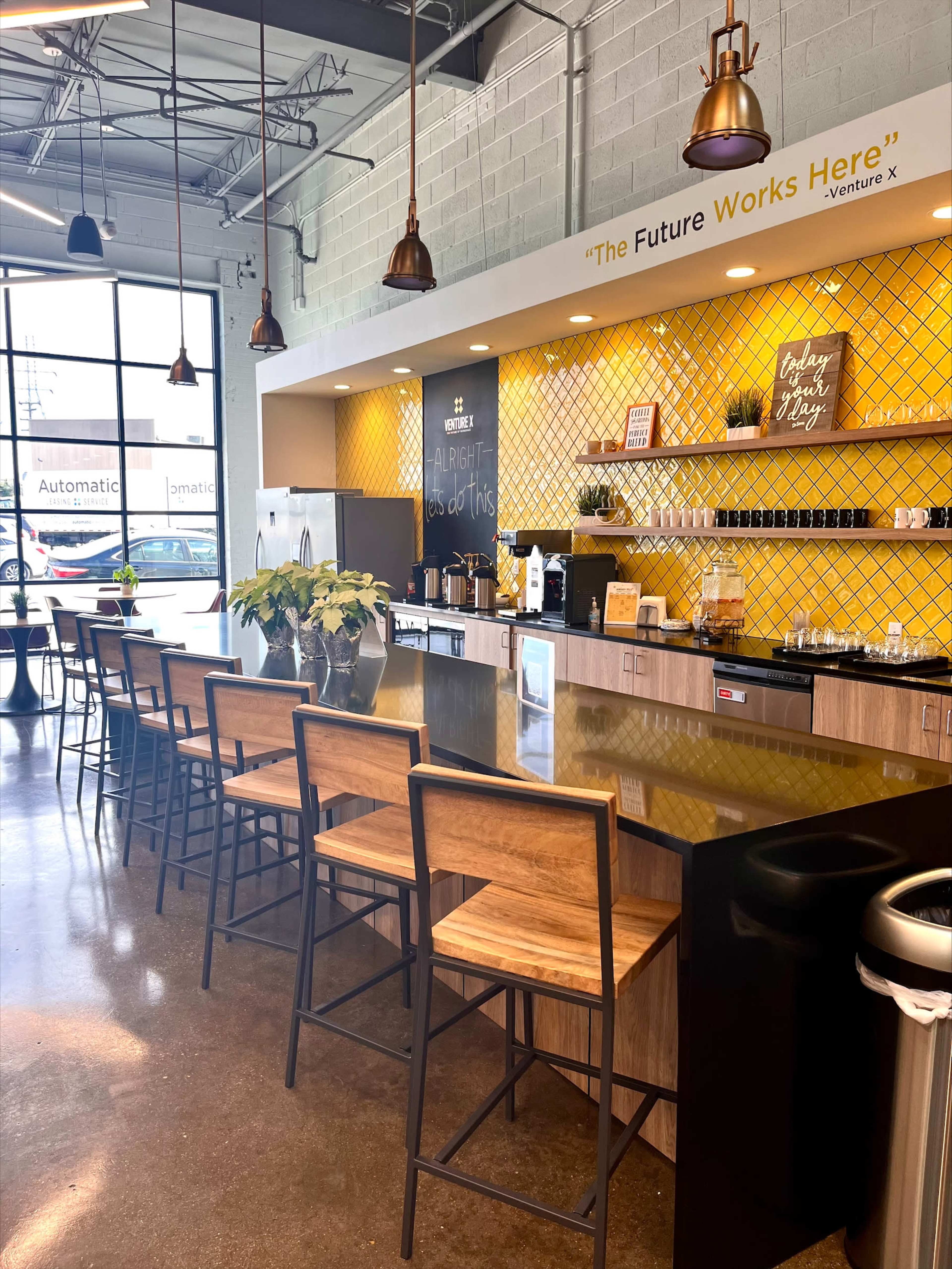 A modern coworking kitchen area with a long wooden countertop, bar stools, and a yellow tiled wall featuring various coffee and kitchen supplies.