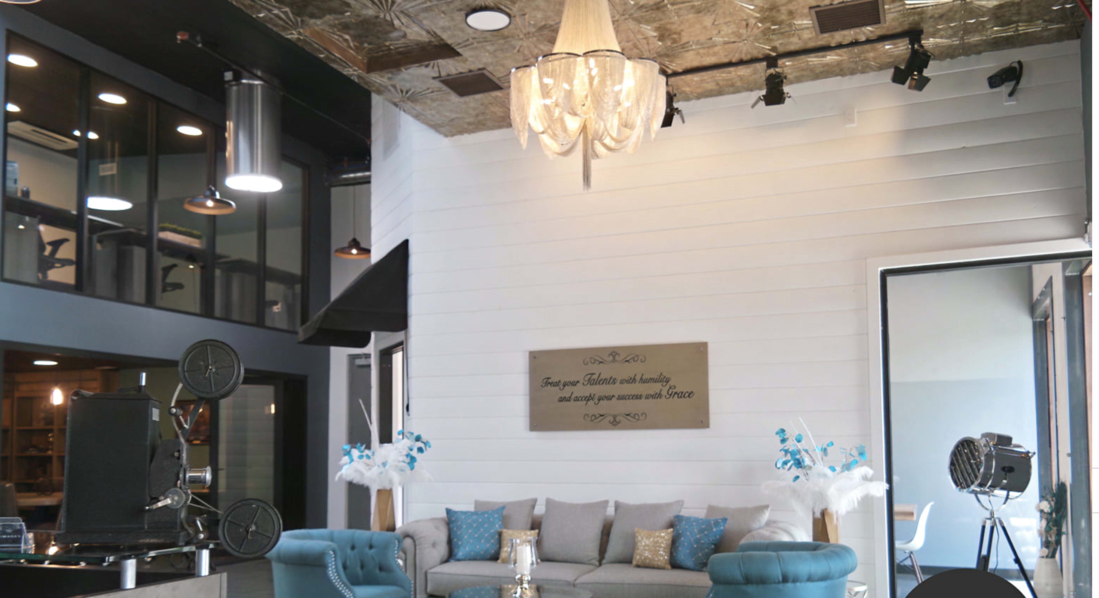 The image shows a modern living space featuring a gray sofa with blue accent pillows, a decorative chandelier, and a sign on the wall that reads a motivational message.