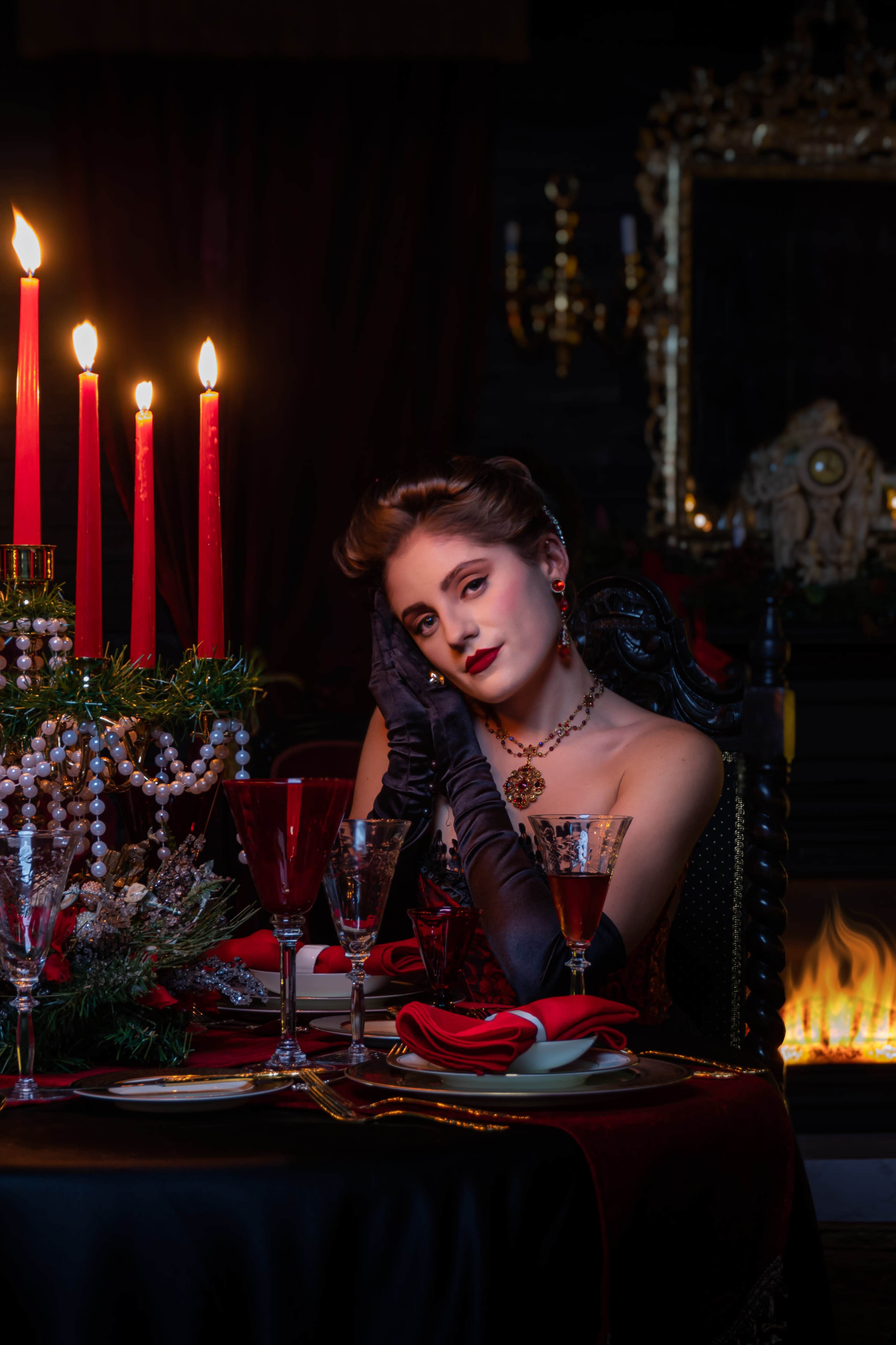 A woman in a red dress sits at a candlelit table, adorned with red candles and elegant glassware, in front of a fireplace.