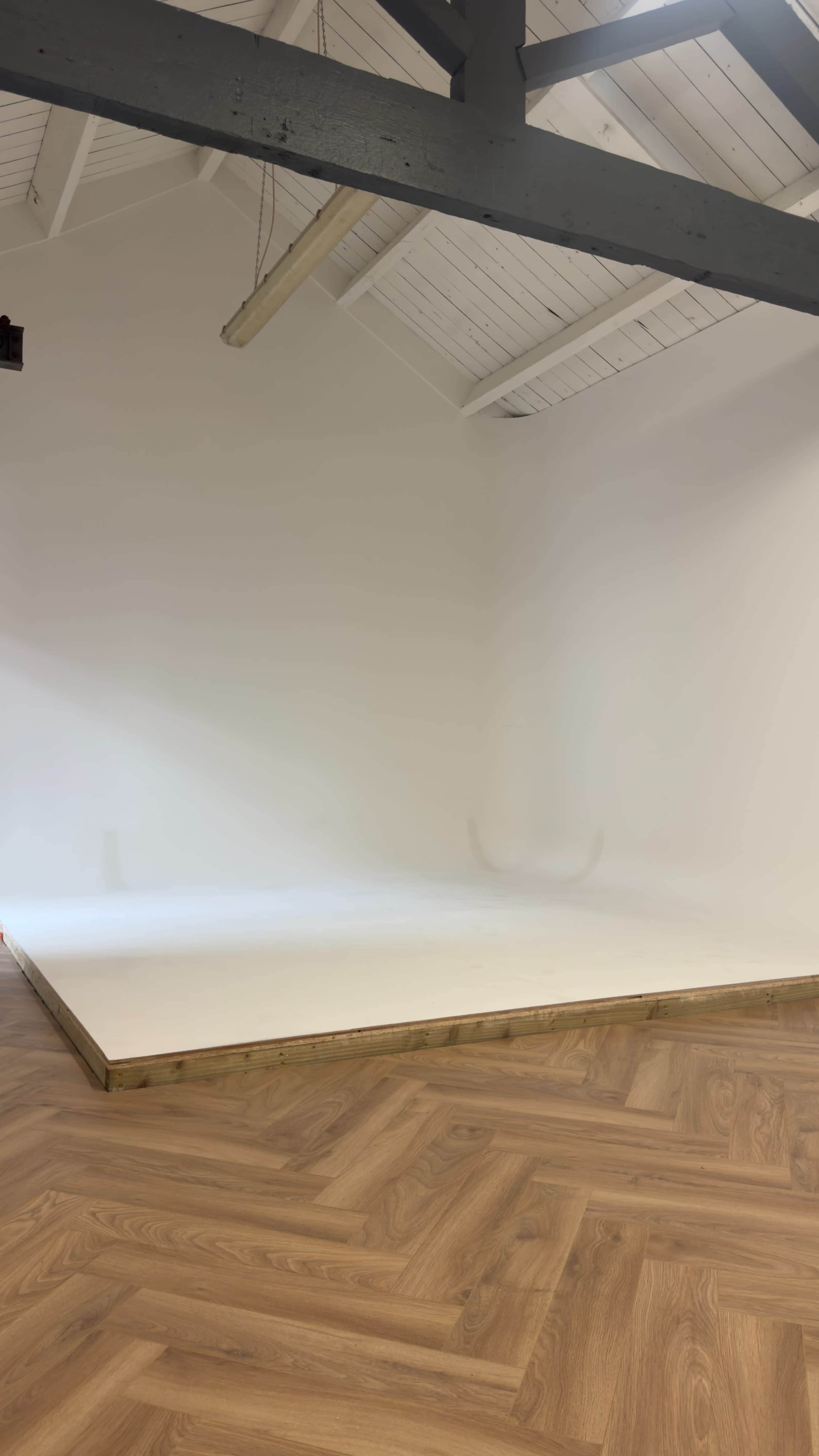 The image shows a spacious, empty room with a wooden floor and a white-walled backdrop setup.