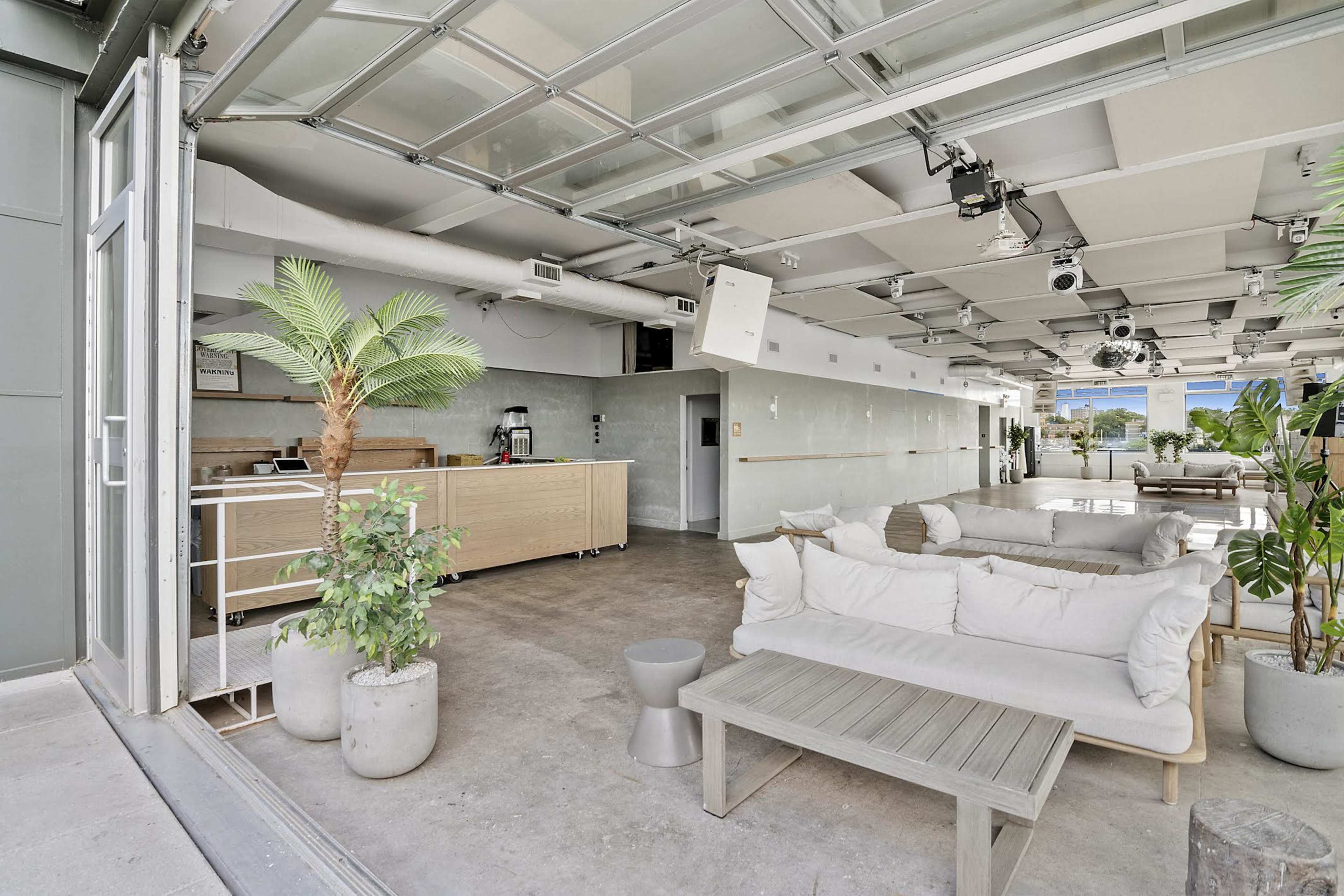 The image shows a modern indoor lounge area with a kitchenette, a white sofa, a wooden coffee table, and tropical plants.