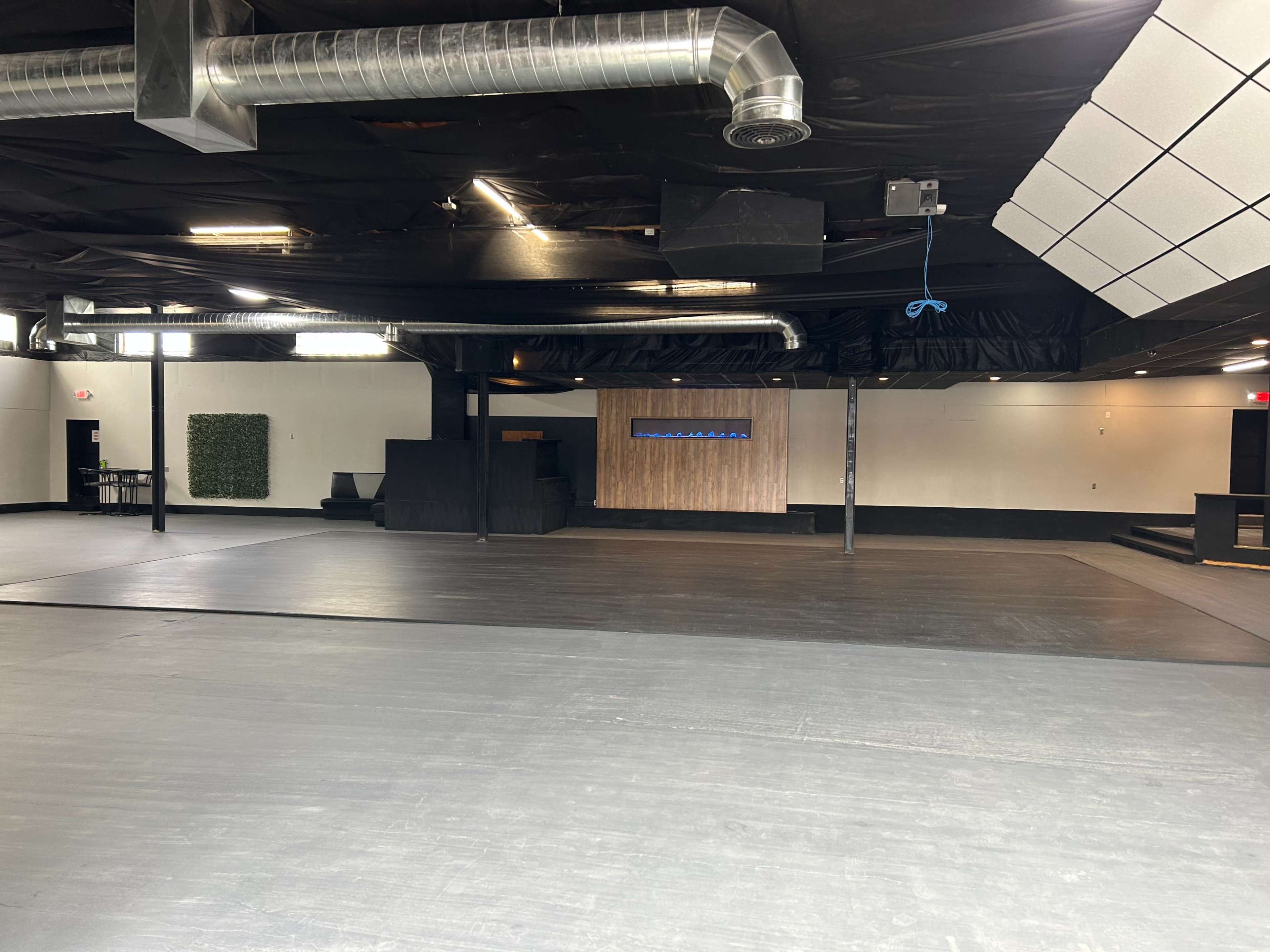 The image shows an empty interior space with a black floor, wooden wall features, and a large area that appears to be designed for events or gatherings.