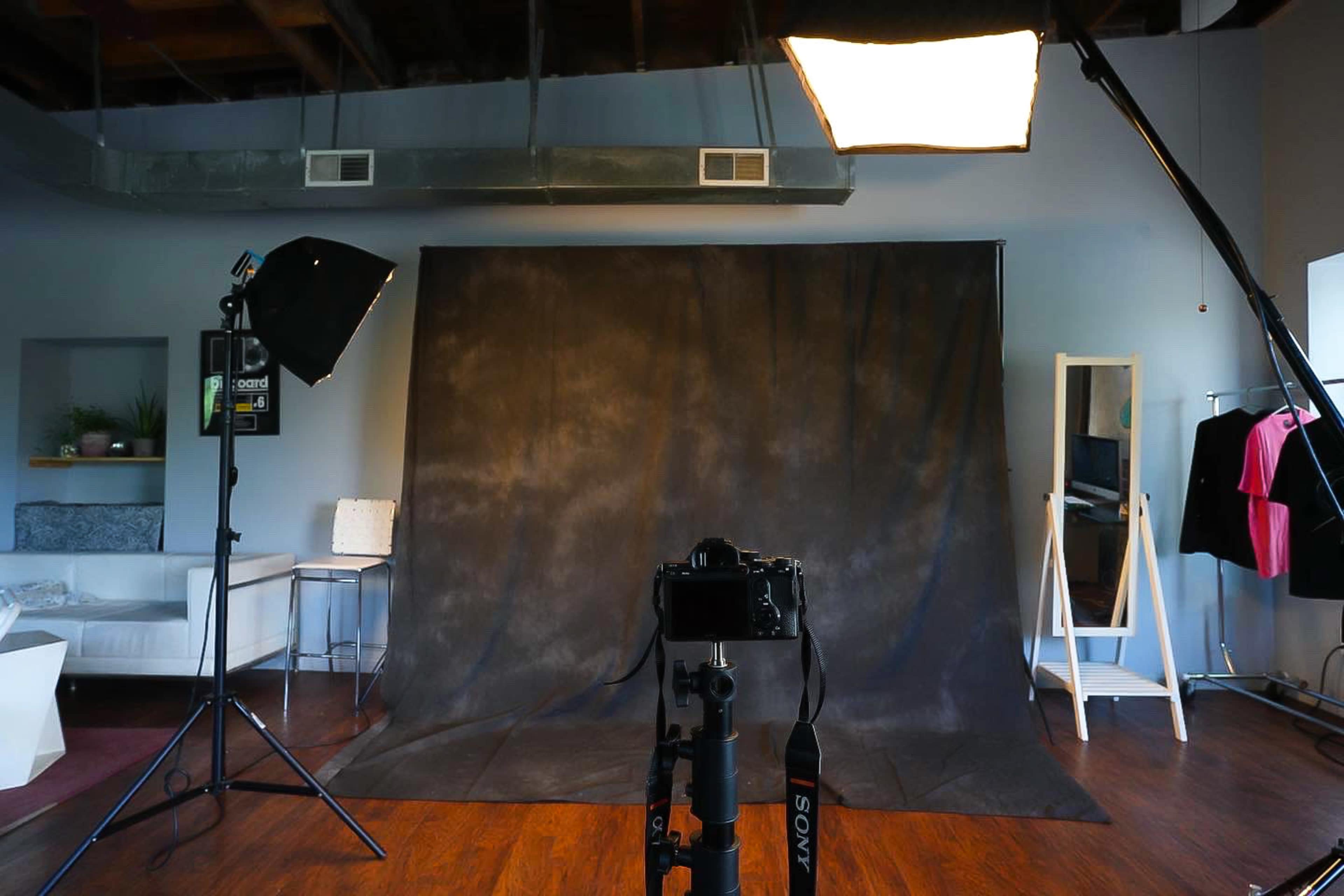 The image shows a photography studio setup with a camera on a tripod, a dark backdrop, lighting equipment, and a few clothing items hanging nearby.