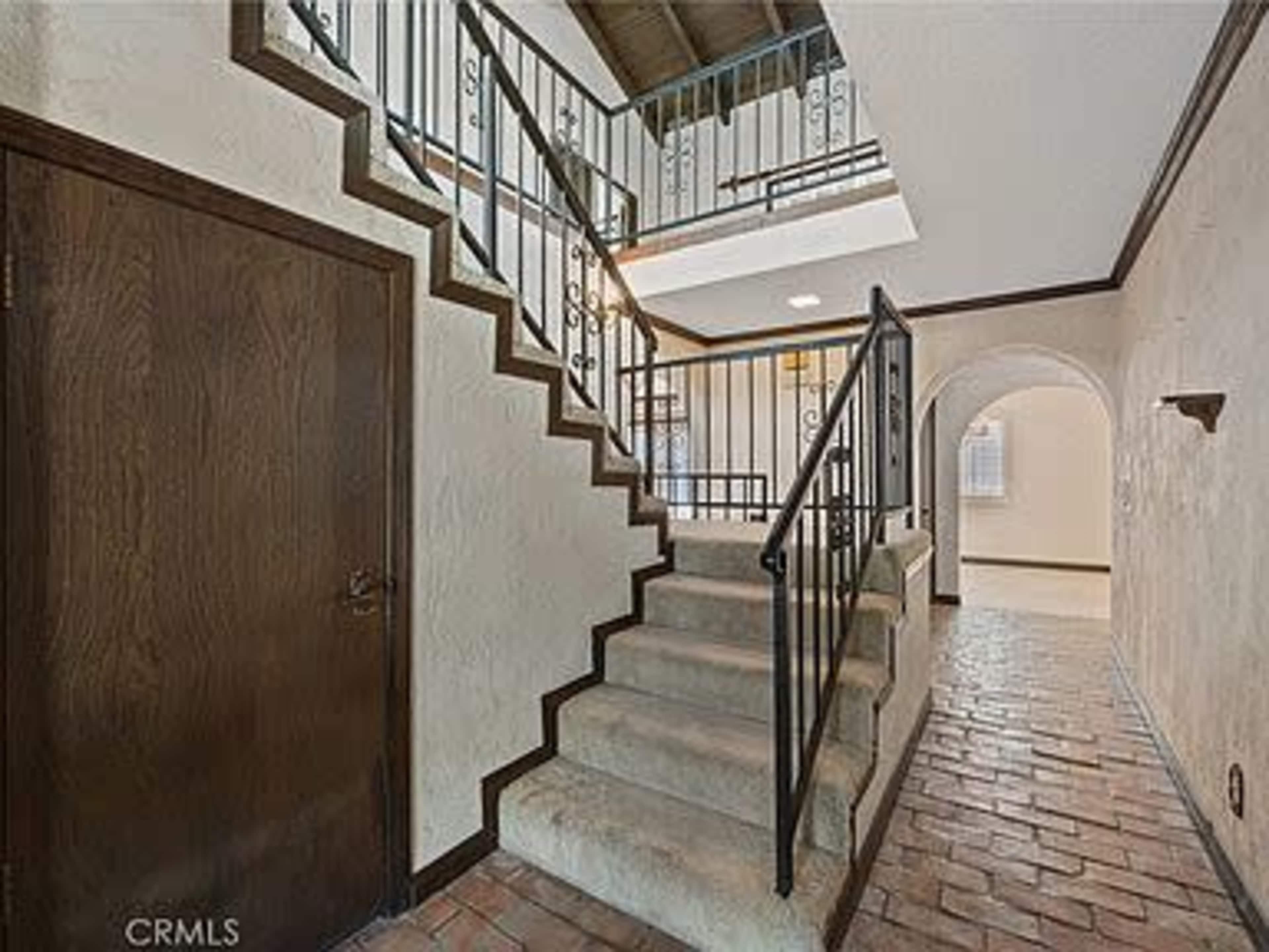 The image shows a staircase with a wrought iron railing, leading to a second floor in a hallway with brick flooring and textured walls.