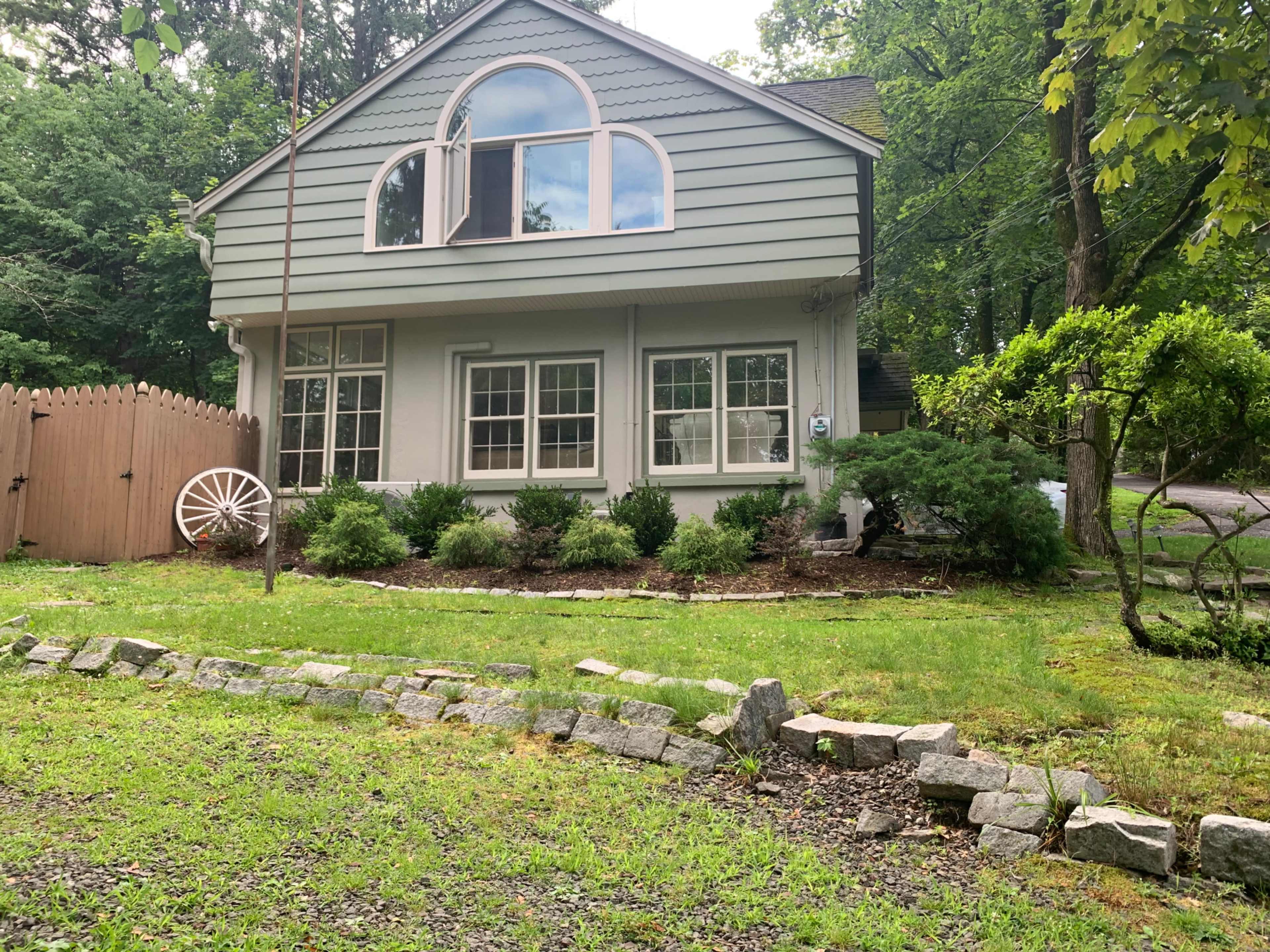 A two-story house with a green exterior and large front windows, surrounded by shrubs and a stone pathway in a wooded area.