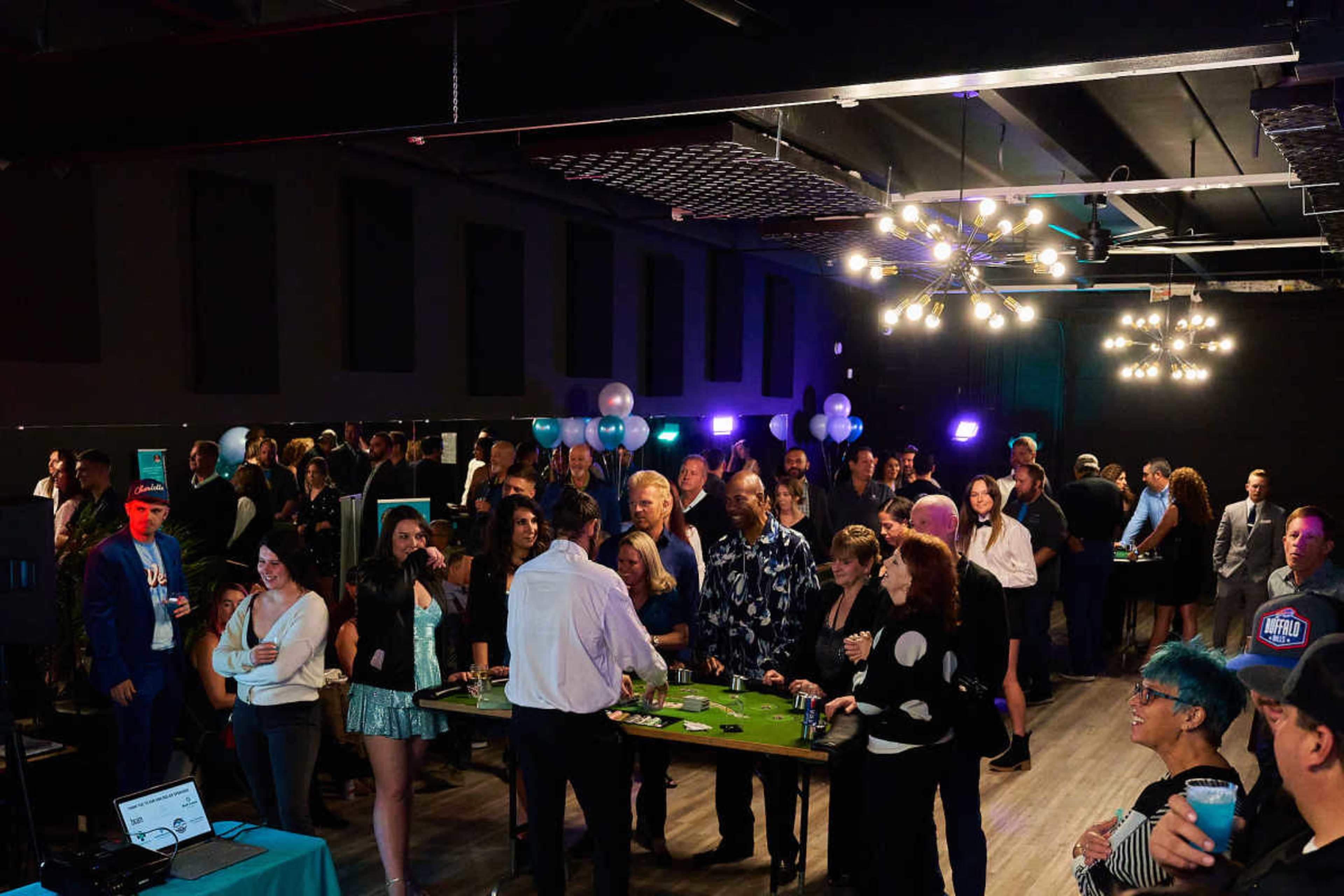 A large group of people gathers in a well-lit venue with a casino-themed setup, including tables for gaming and decorations.