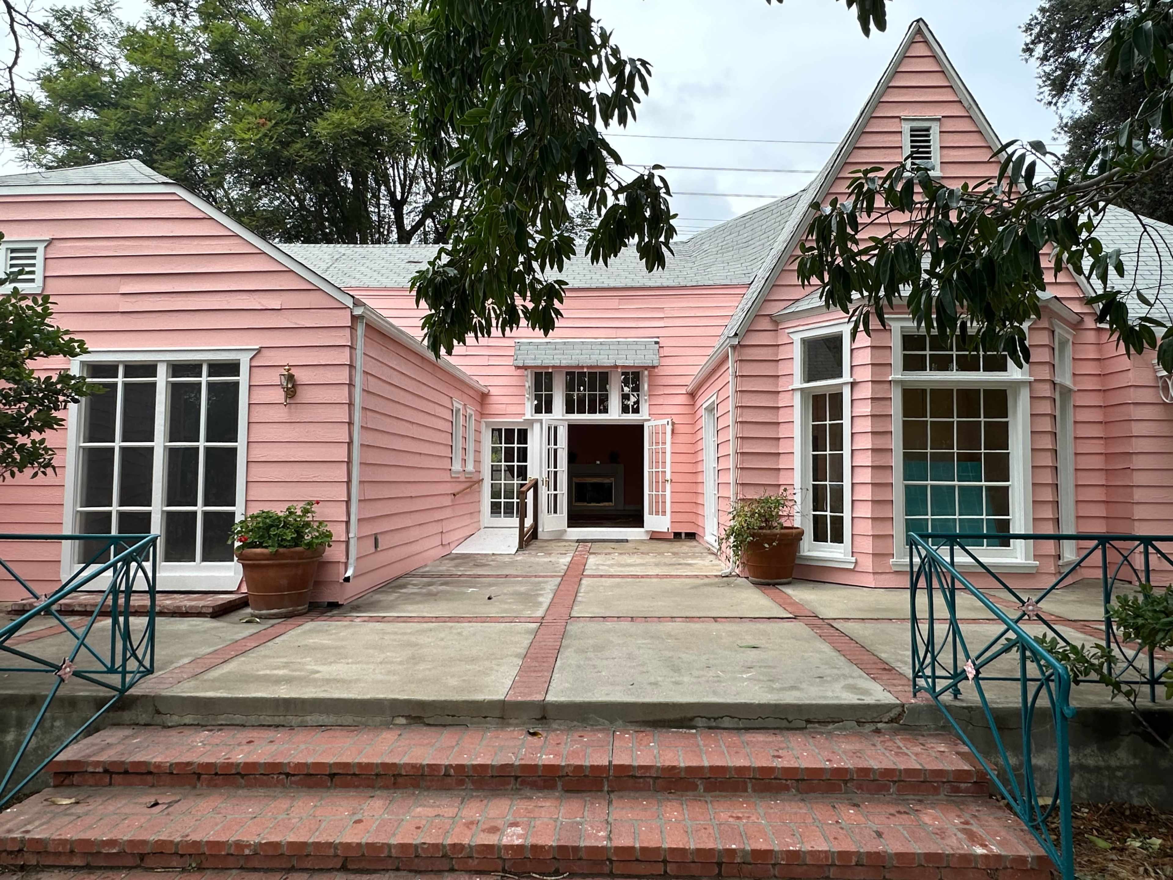 A pink house with a triangular roof, large windows, and a brick pathway leading to its entrance.