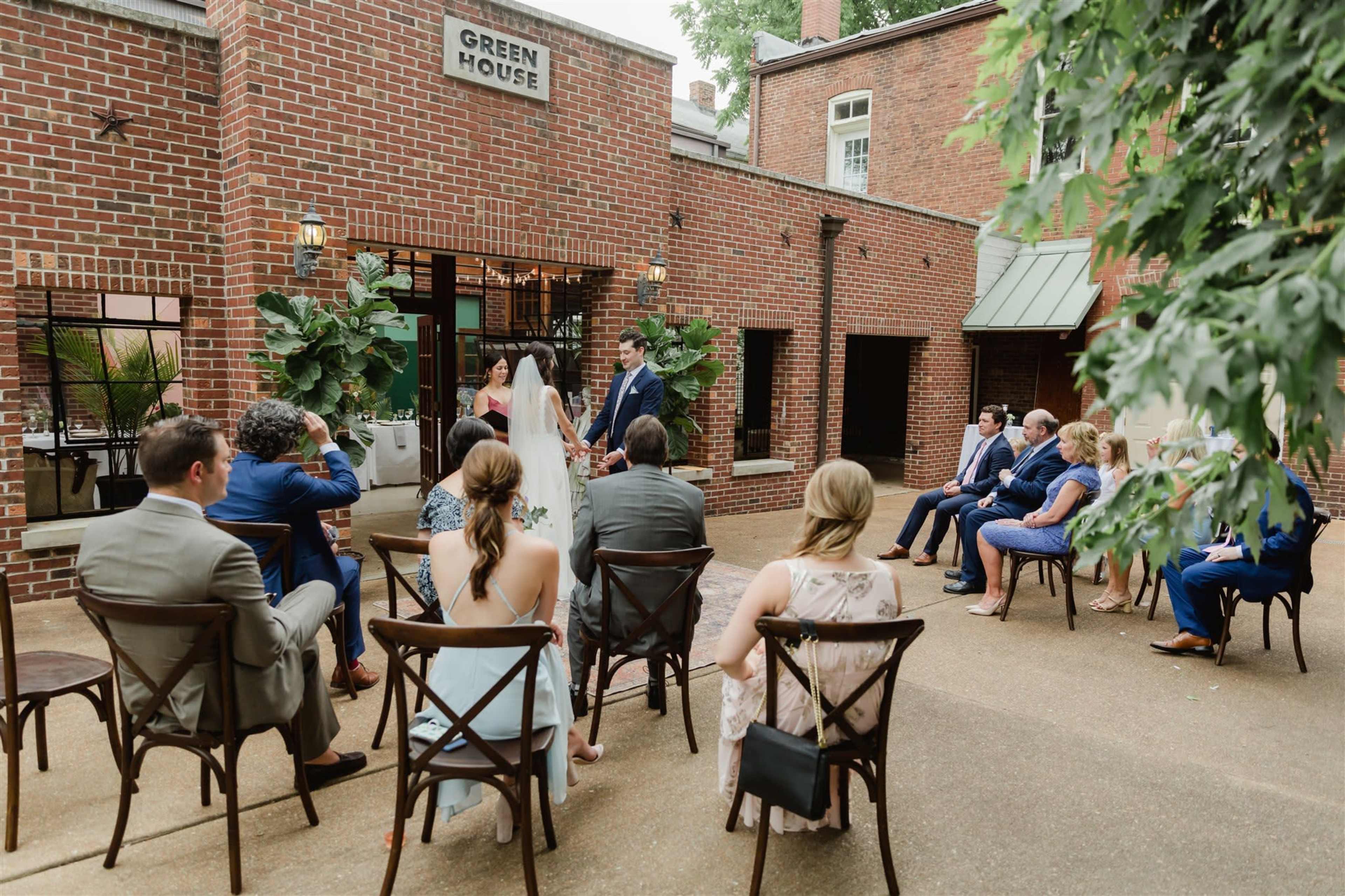 A couple stands at the front of a courtyard, exchanging vows while guests seated in rows of chairs watch.