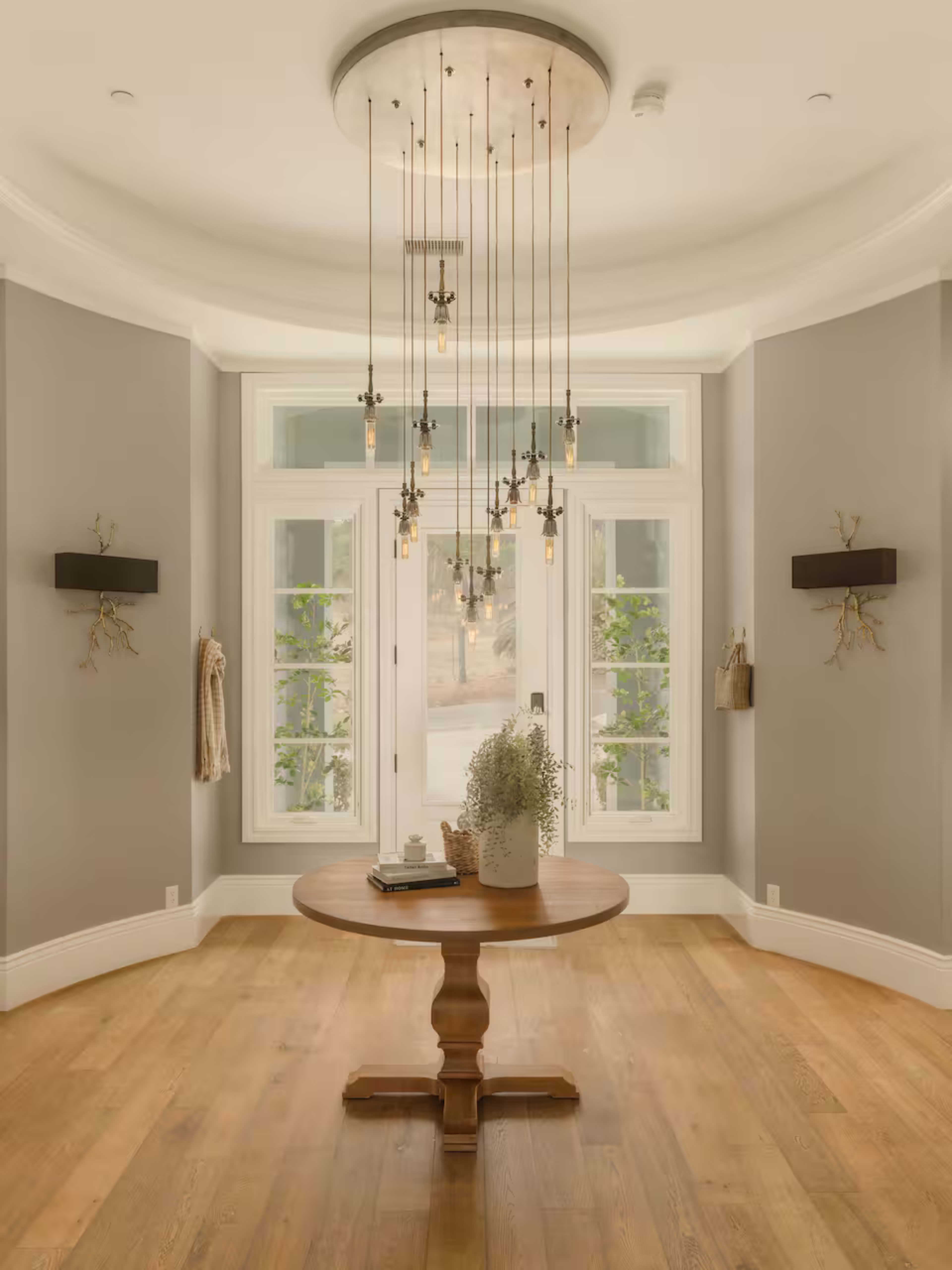 The image features a spacious room with a round wooden table in the center, illuminated by a modern chandelier, and large windows that provide natural light.