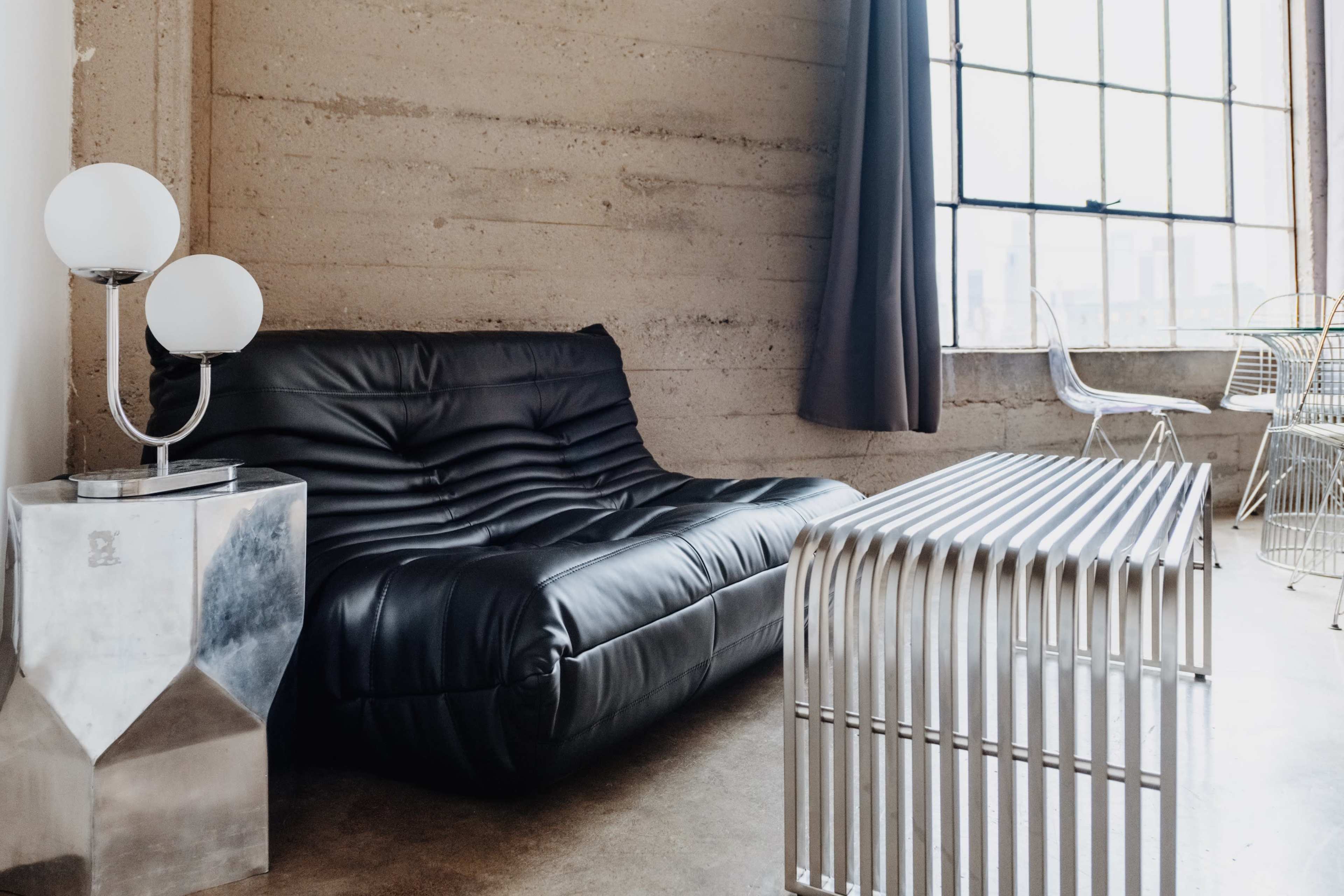 A modern living space featuring a black sofa, a metallic side table, and a glass window with sheer curtains.